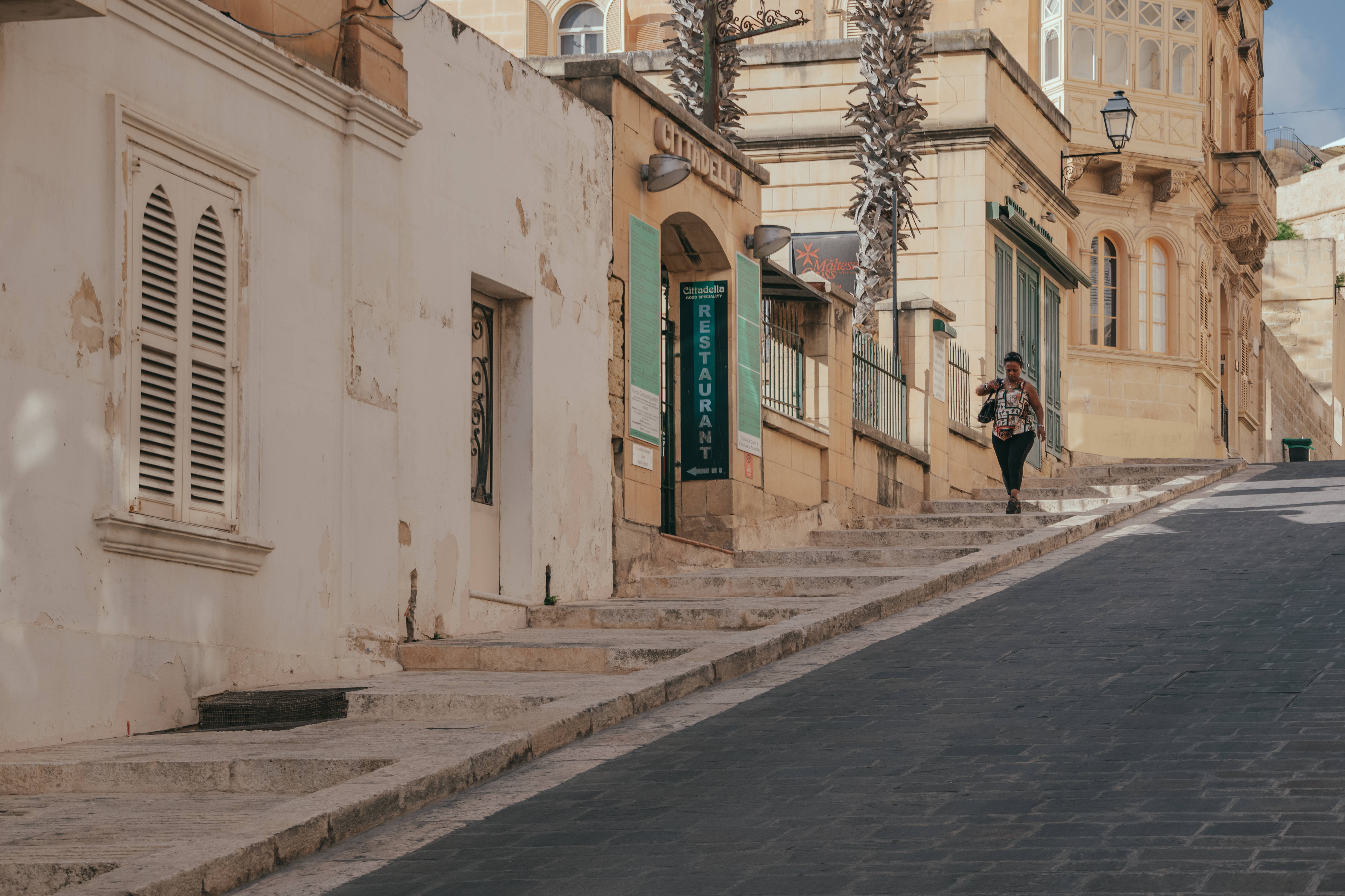 Malta 桌布 壁紙 — 一名女子沿著一條陽光明媚的狹窄街道走上石階，街道兩旁是陳舊的米色建築和棕櫚樹。