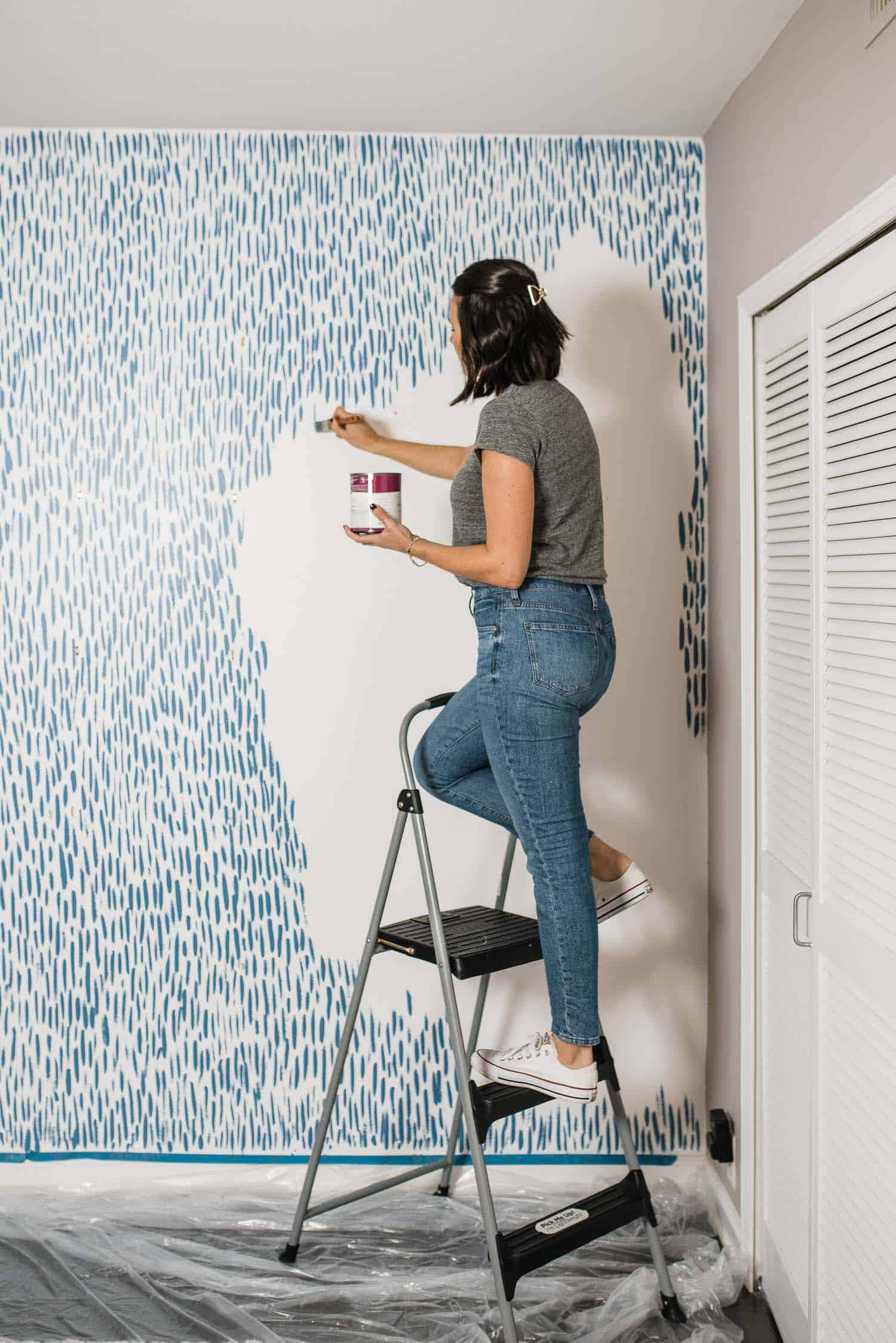 Wall Painting 桌布 壁紙 — 一名女子站在梯子上，在一個地板鋪有塑膠布的房間裡，在牆上畫藍色抽象圖案。