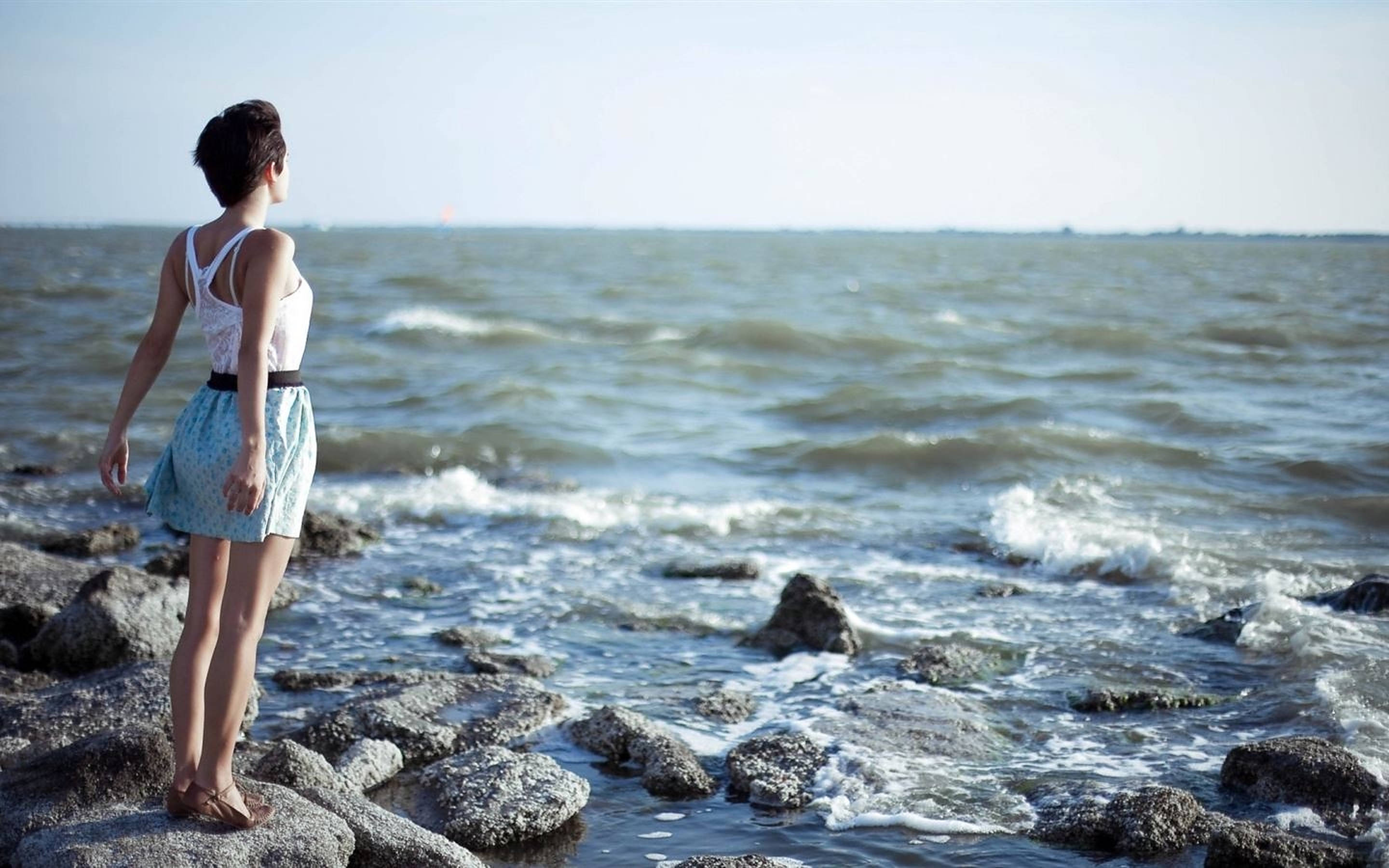 Breathing 桌布 壁紙 — 一名女子赤腳站在岩石海岸上，在明亮的日光下凝視著廣闊的海洋。