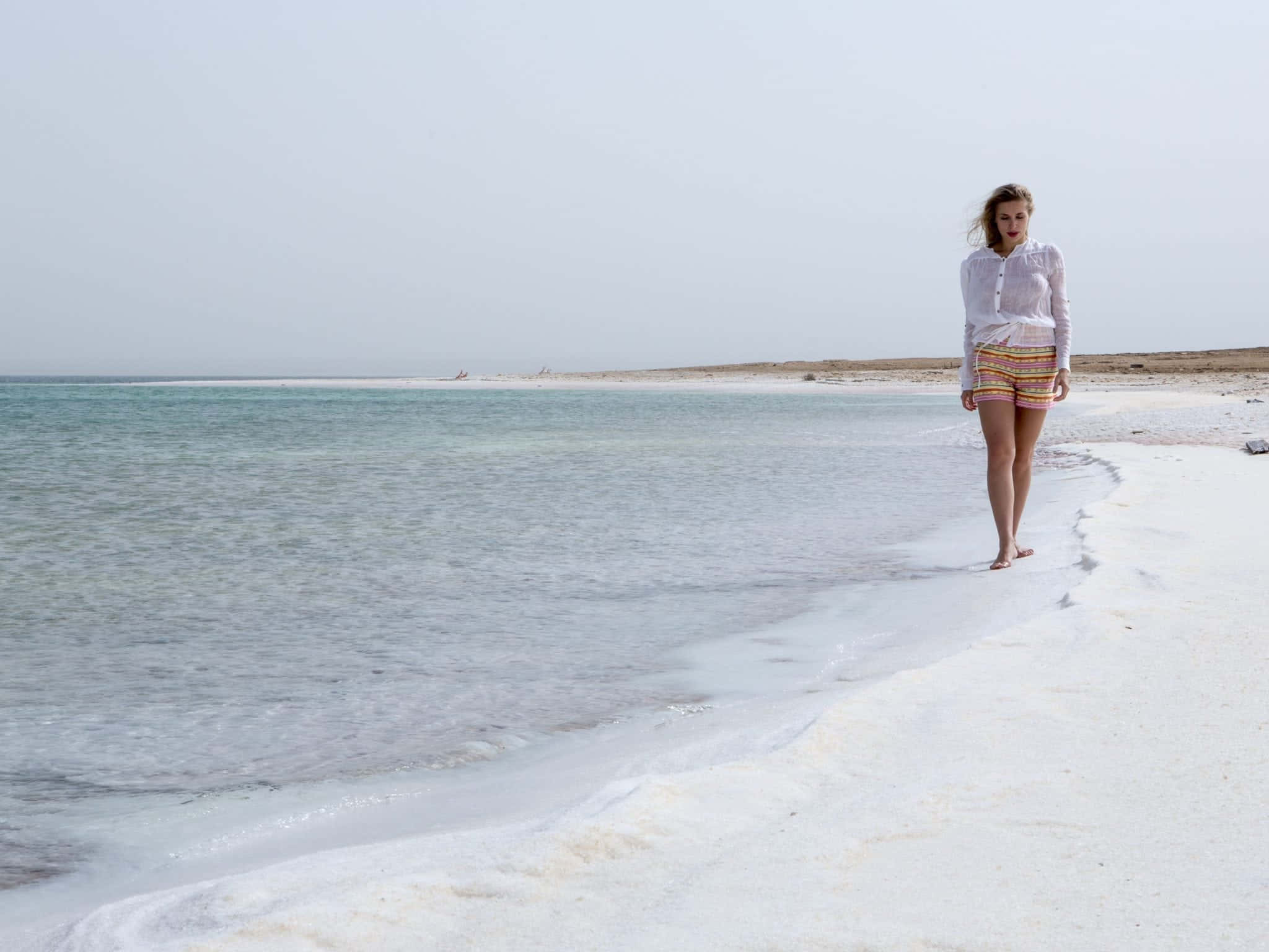 Israel 桌布 壁紙 — 一名女子赤腳走在超現實的白色鹽灘上，在朦朧的天空下，碧綠的海水輕拍著海岸。