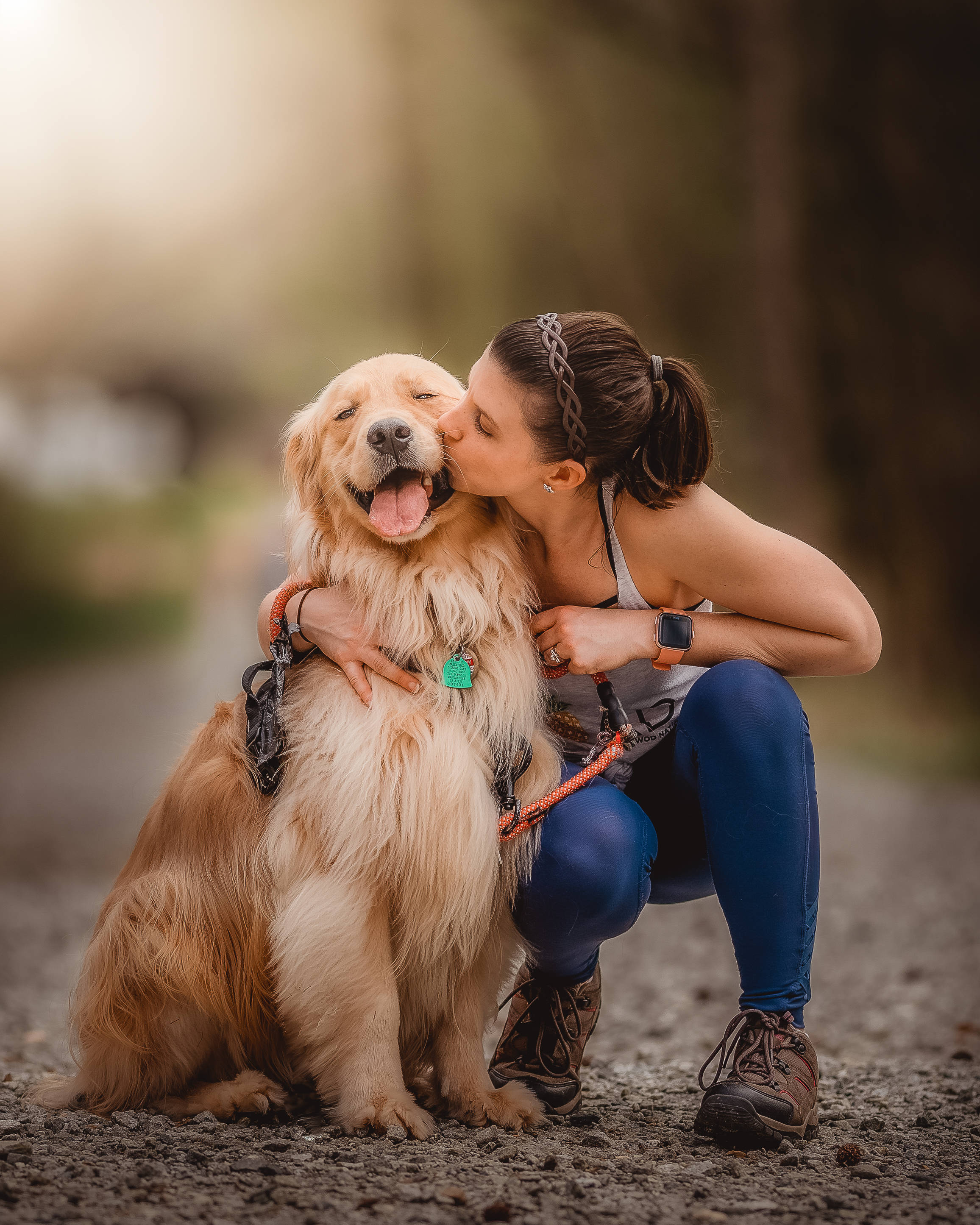 Dog And Girl 桌布 壁紙 — 一名女子跪下來親吻一隻黃金獵犬的臉頰，他們一起坐在陽光普照的森林中的碎石路上。