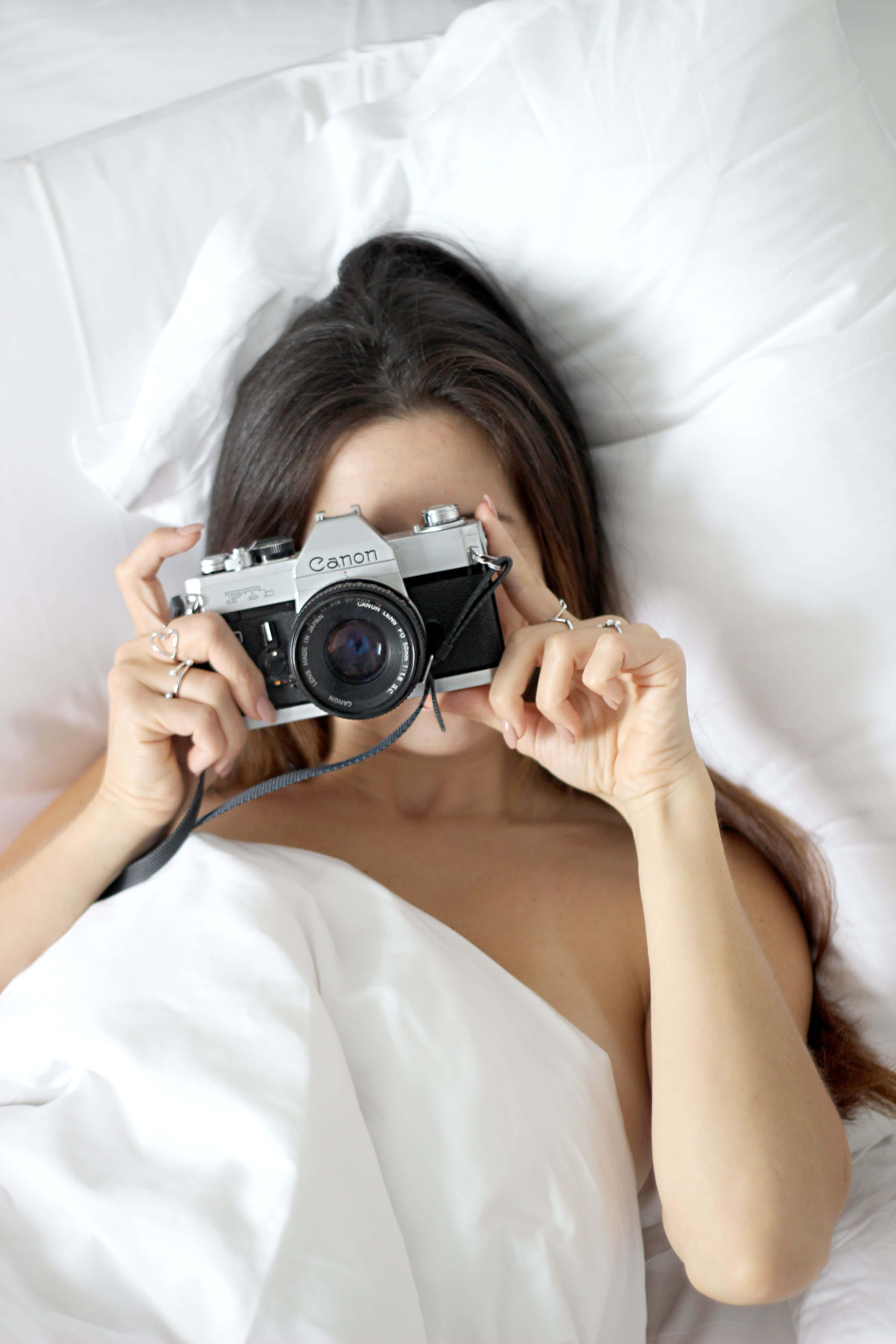 Holding Camera 桌布 壁紙 — 一名女子躺在白色床上，將一部復古Canon相機舉到臉前，部分遮擋了她的視線。