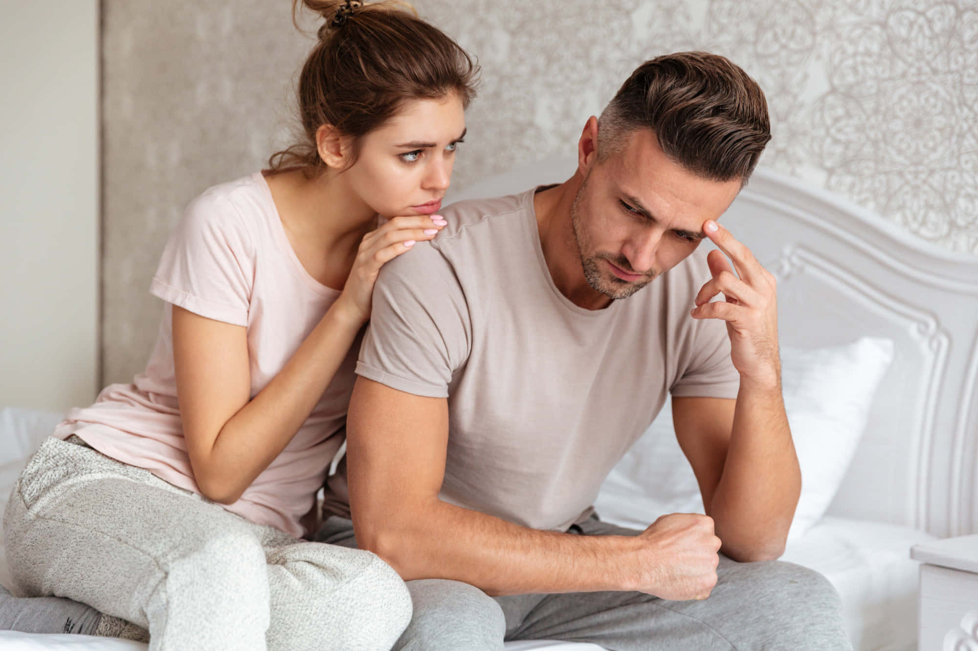 couple in bed 桌布 壁紙 — 一名女子靠近坐在床上的男子，她的手放在他的肩膀上，而他則在痛苦地揉著太陽穴。
