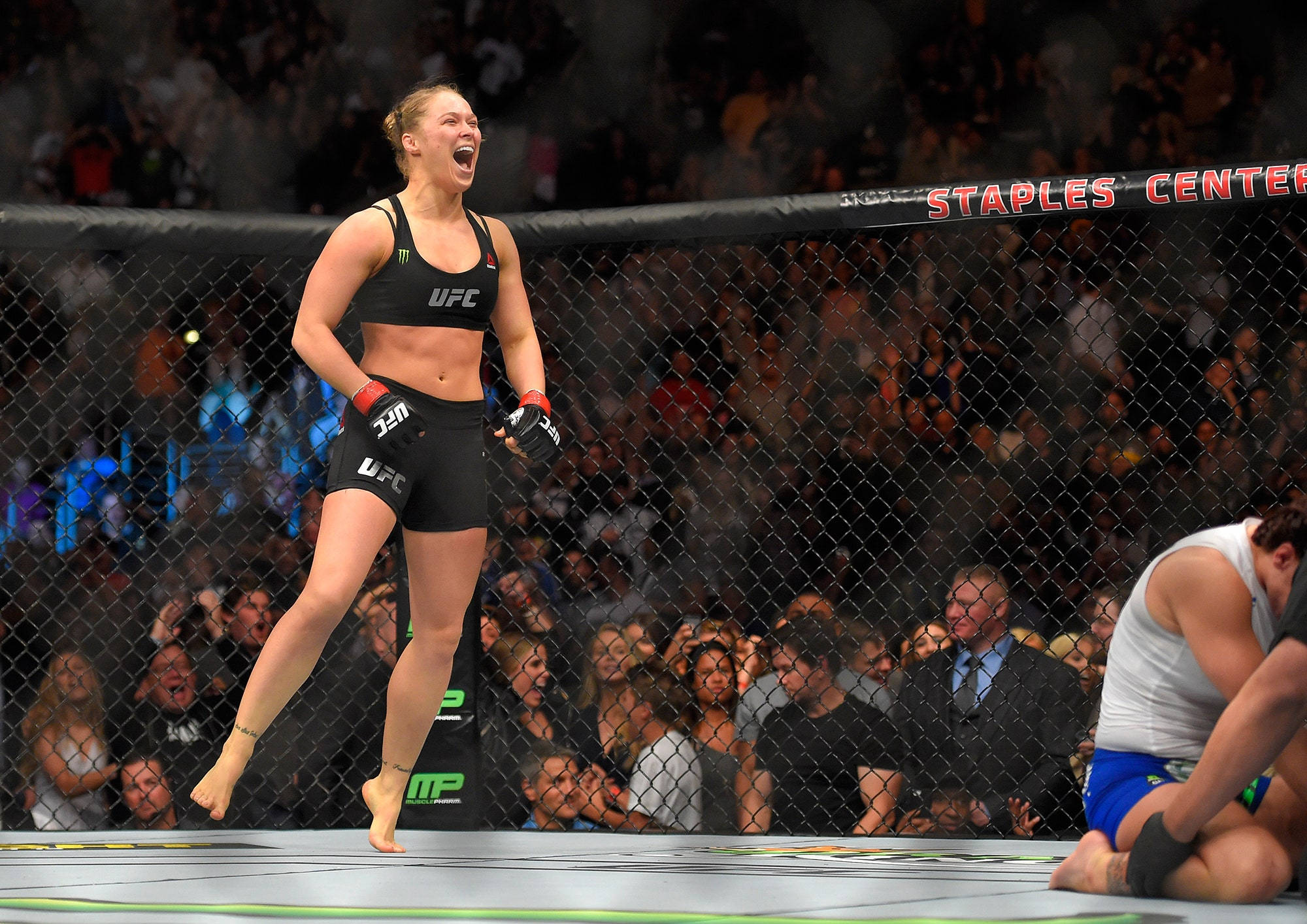 Rousey 桌布 壁紙 — 一名女性UFC拳手在八角籠中慶祝勝利，對手跪地敗北，觀眾歡呼雷動。