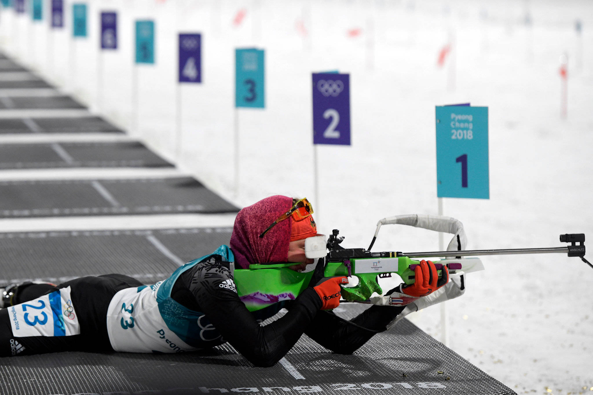 運動 桌布 壁紙 — 一名女性冬季兩項運動員穿著鮮豔裝備，俯臥在雪地上，用步槍瞄準帶有奧運五環和平昌2018標誌的靶標。