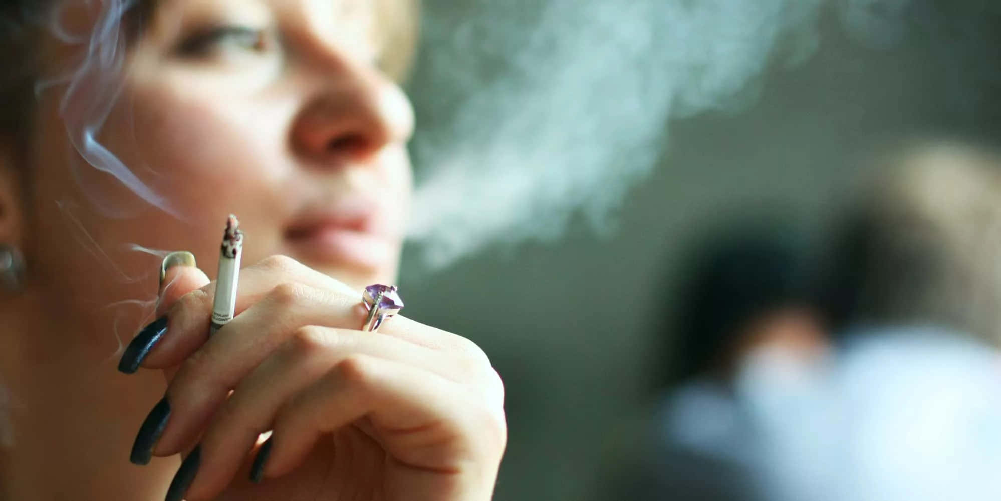 cigarettes 桌布 壁紙 — 一名女性呼出煙霧，手中拿著一支香煙，手指塗有深色指甲油並戴著紫色戒指。