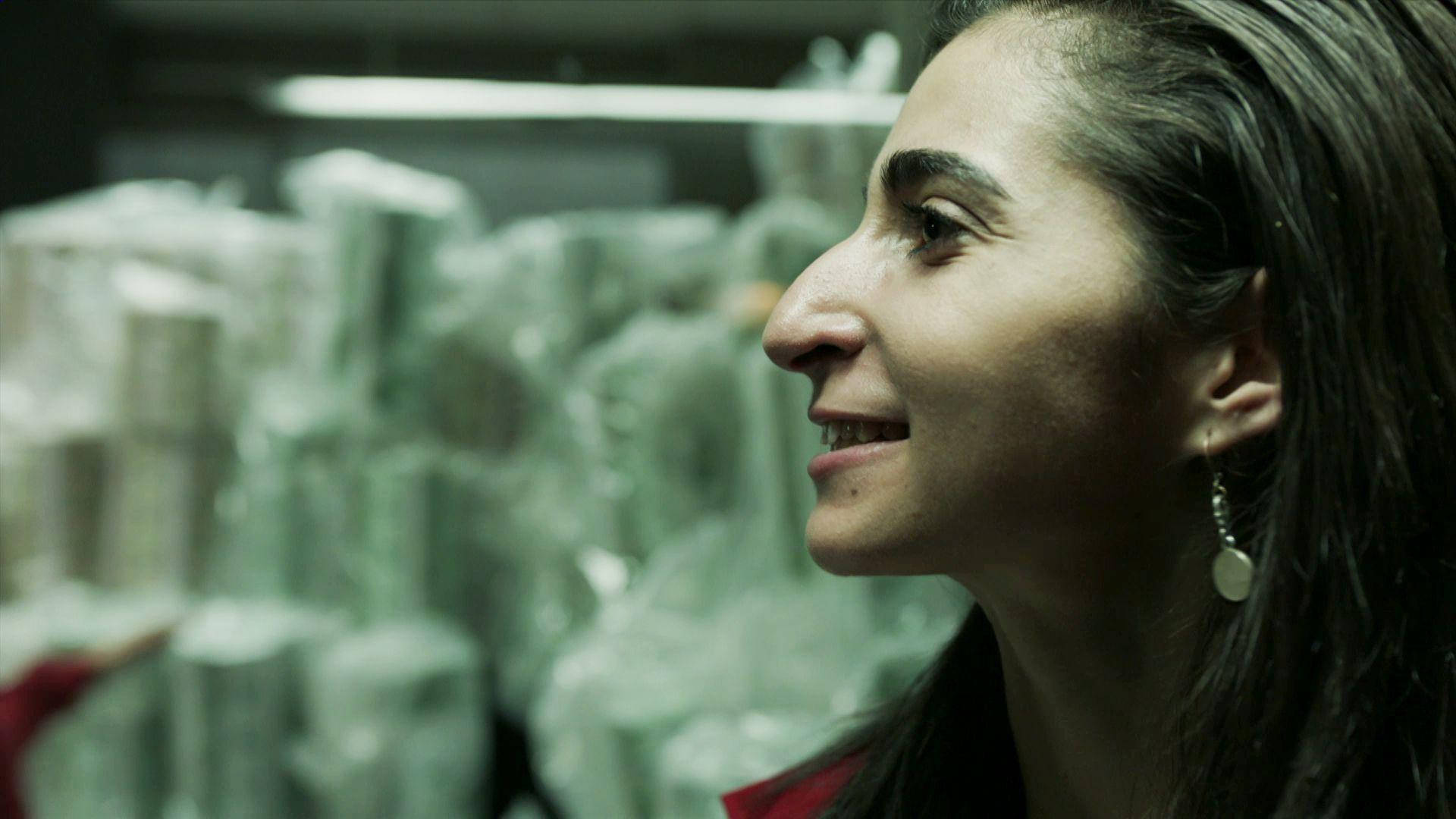 nairobi money heist 桌布 壁紙 — 一名女性在冷色螢光燈下，以模糊的塑膠包裝堆疊物品為背景，側面微笑。