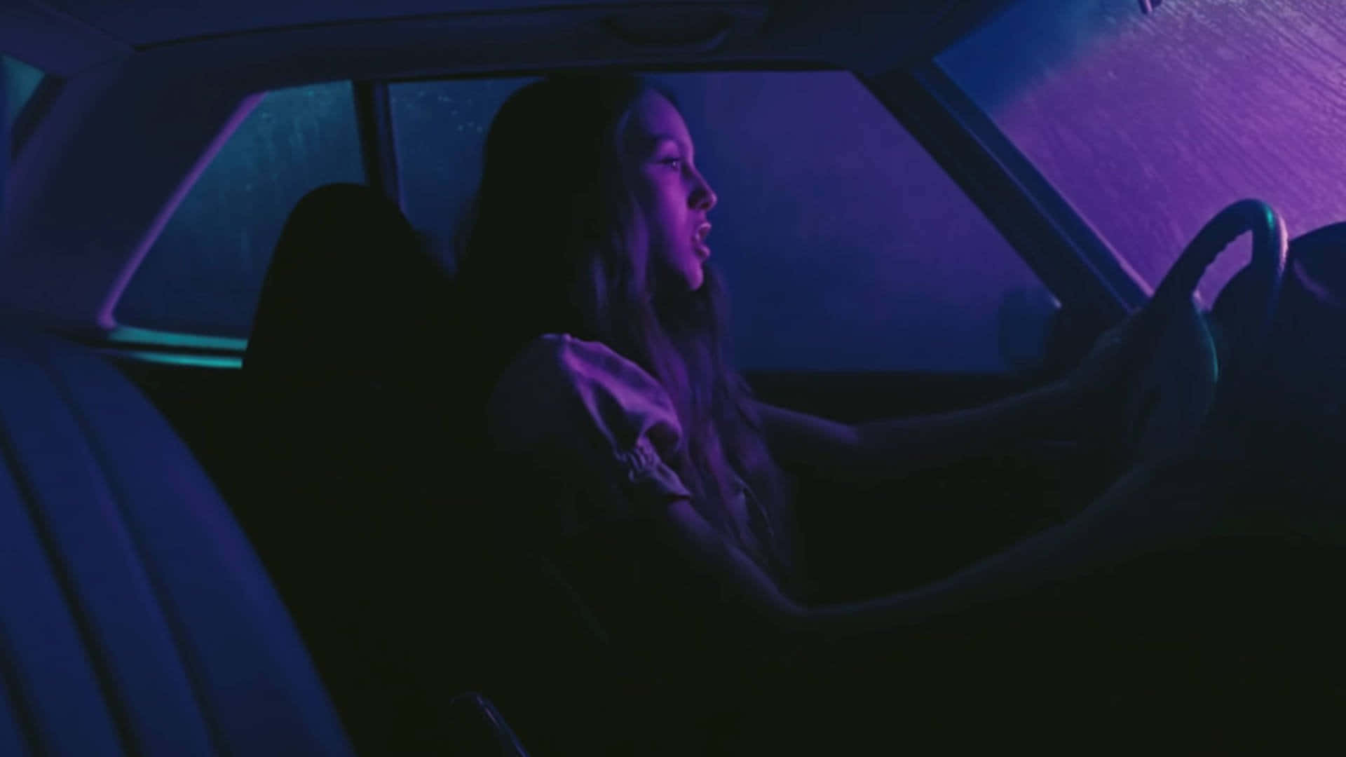 奧莉維亞·羅德里戈 桌布 壁紙 — 一名女性在夜間駕駛汽車，車內被街道標誌的霓虹紫色和藍色燈光籠罩。