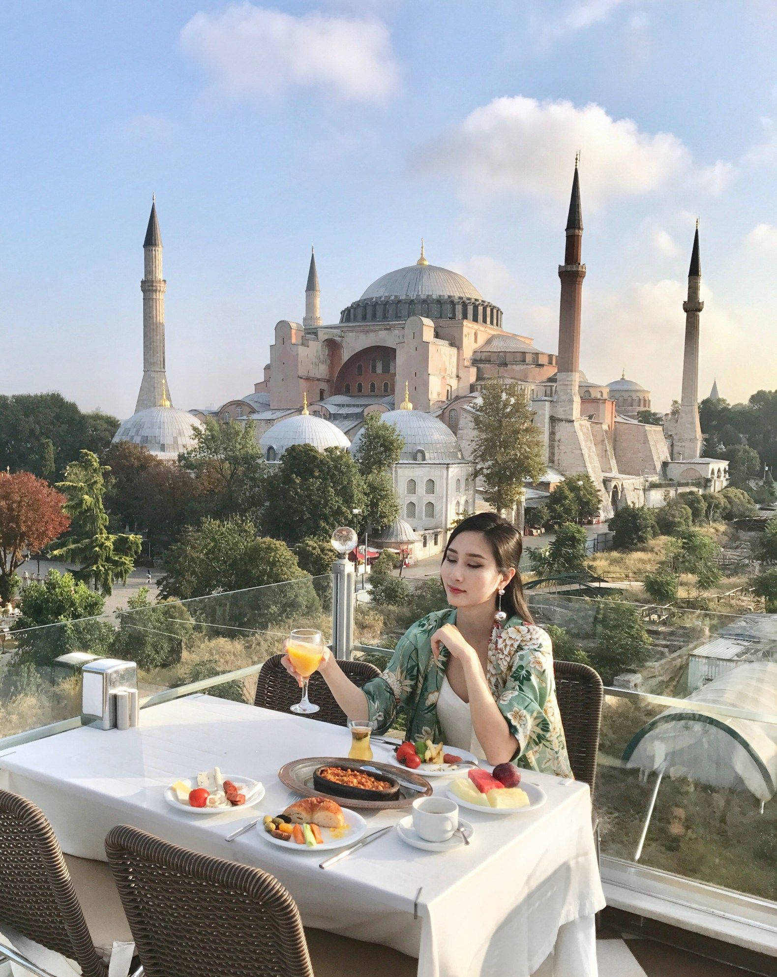 Istanbul 桌布 壁紙 — 一名女性在屋頂露臺上享用早餐，欣賞著藍天白雲下聖索菲亞大教堂的壯麗景色。