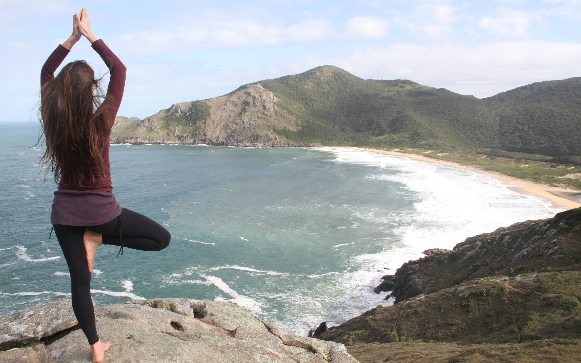 Yoga 桌布 壁紙 — 一名女性在懸崖上以樹式平衡，俯瞰著碧綠色的海洋、沙灘和連綿起伏的綠色山丘。