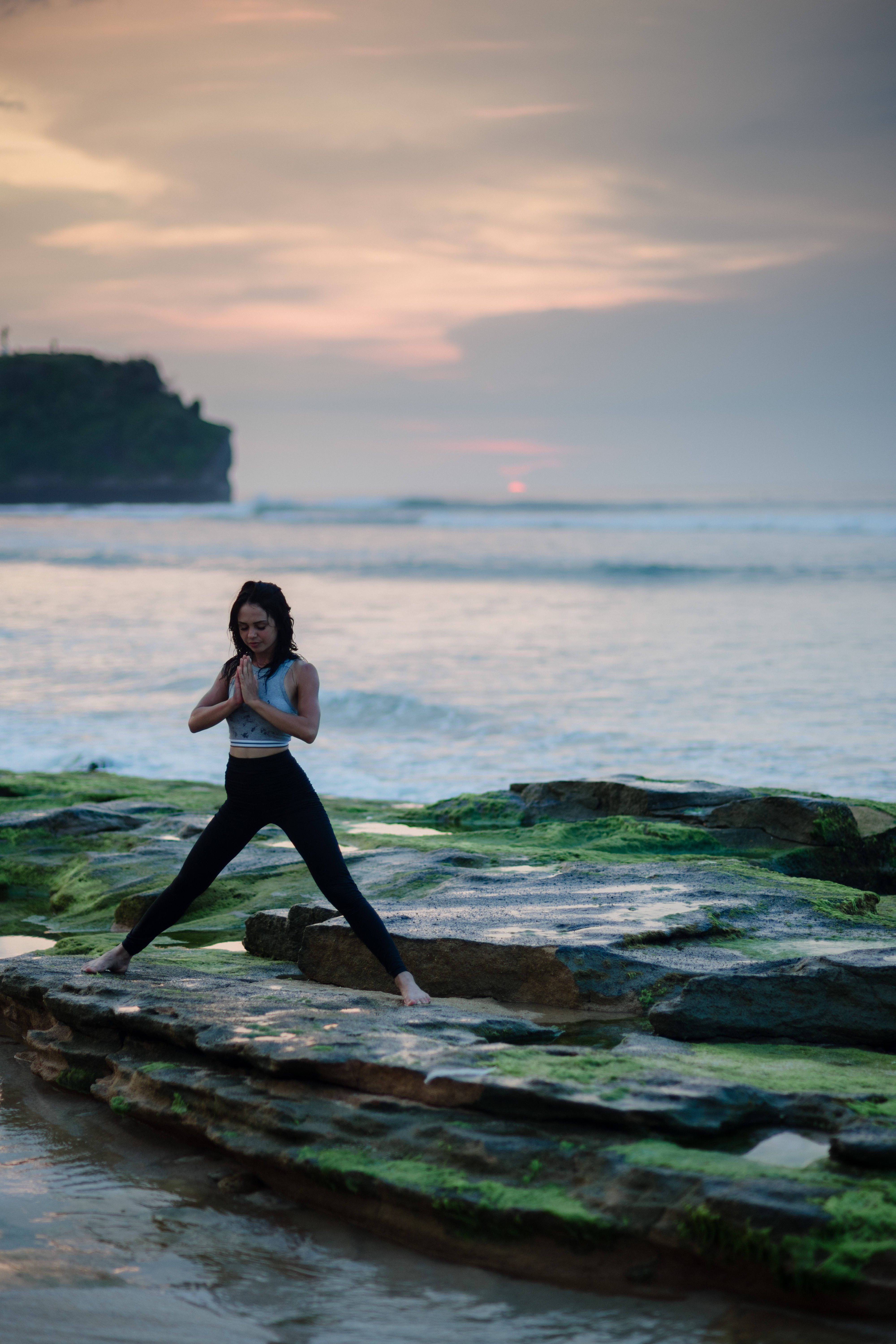 Yoga 桌布 壁紙 — 一名女性在日落時分，於海邊長滿苔蘚的岩石上以寬闊姿勢練習瑜伽，遠處有懸崖。