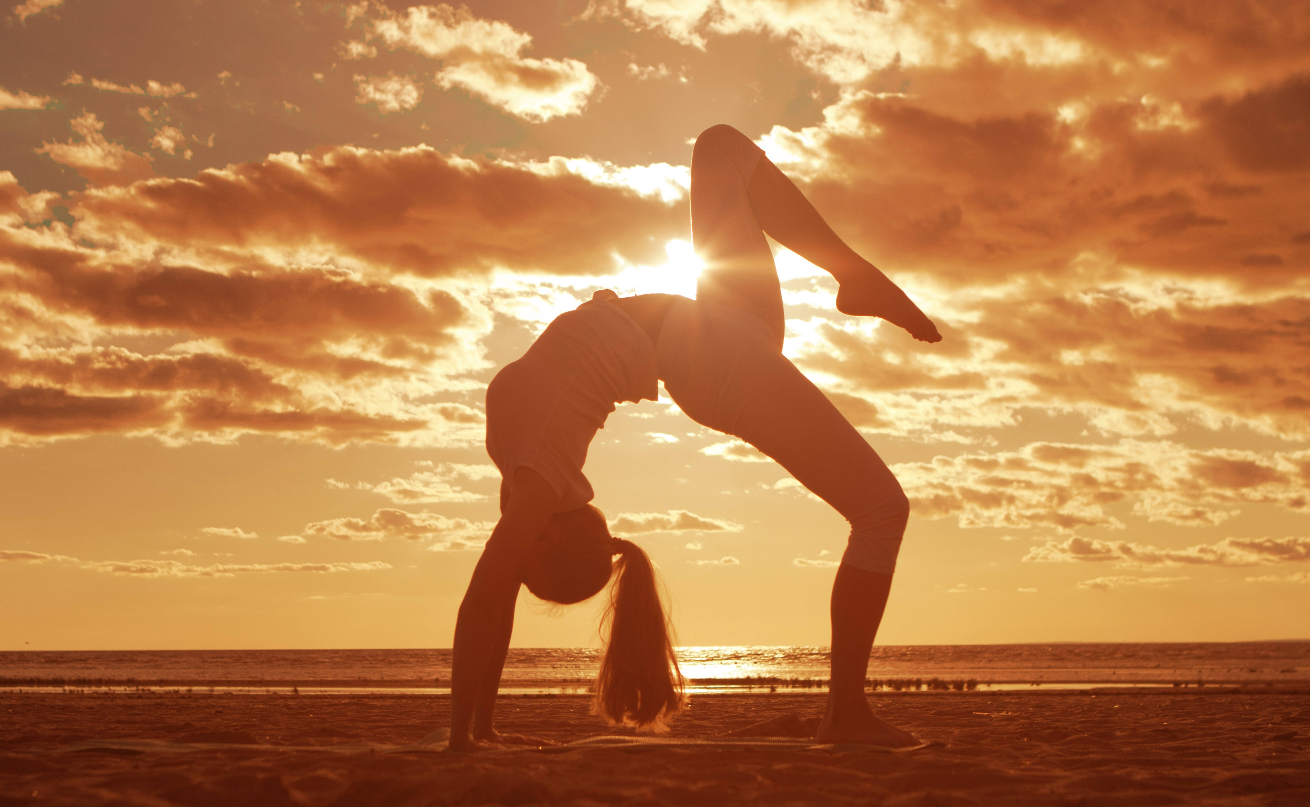 Yoga 桌布 壁紙 — 一名女性在日落時分的沙灘上，以金色雲彩和海洋為背景，優雅地做出後彎動作。