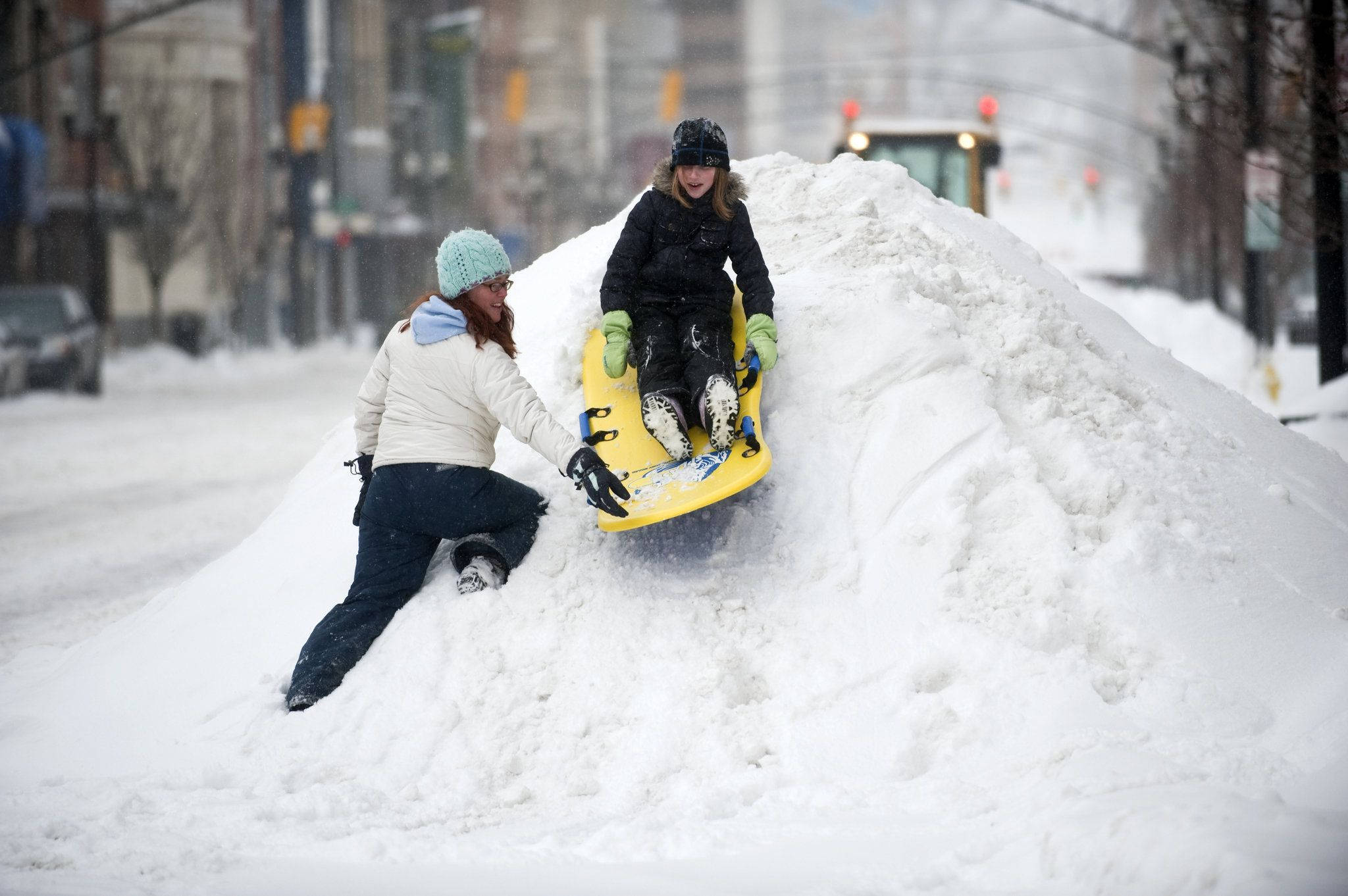 雪 桌布 壁紙 — 一名女性在積雪的市區街道上，推著一個坐在黃色雪橇上的小孩，位於大雪堆的頂端。