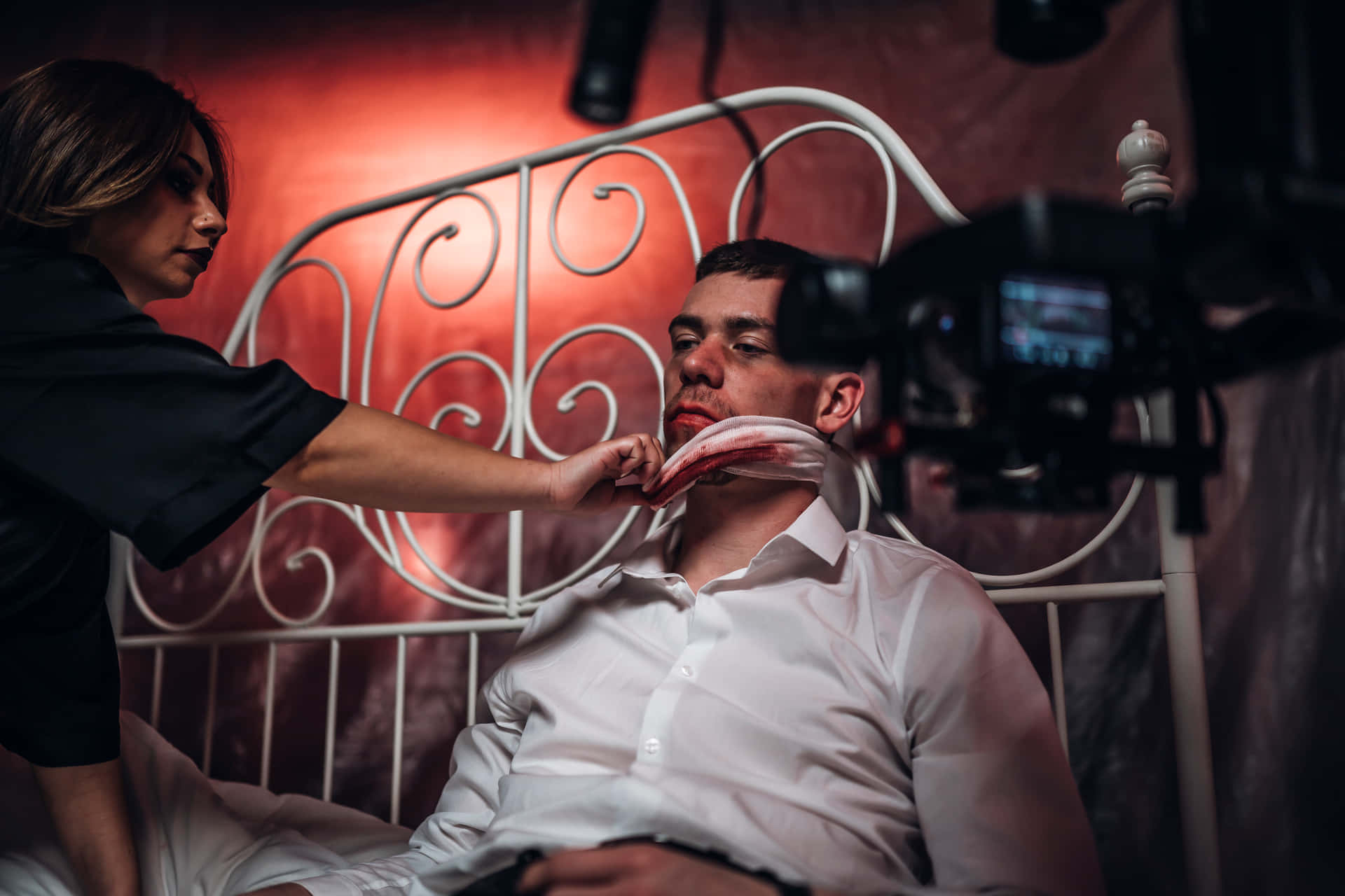 acting 桌布 壁紙 — 一名女性在紅色燈光下，於一張帶有華麗白色床頭板的床上，將假血塗在男性脖子上並拍攝他。