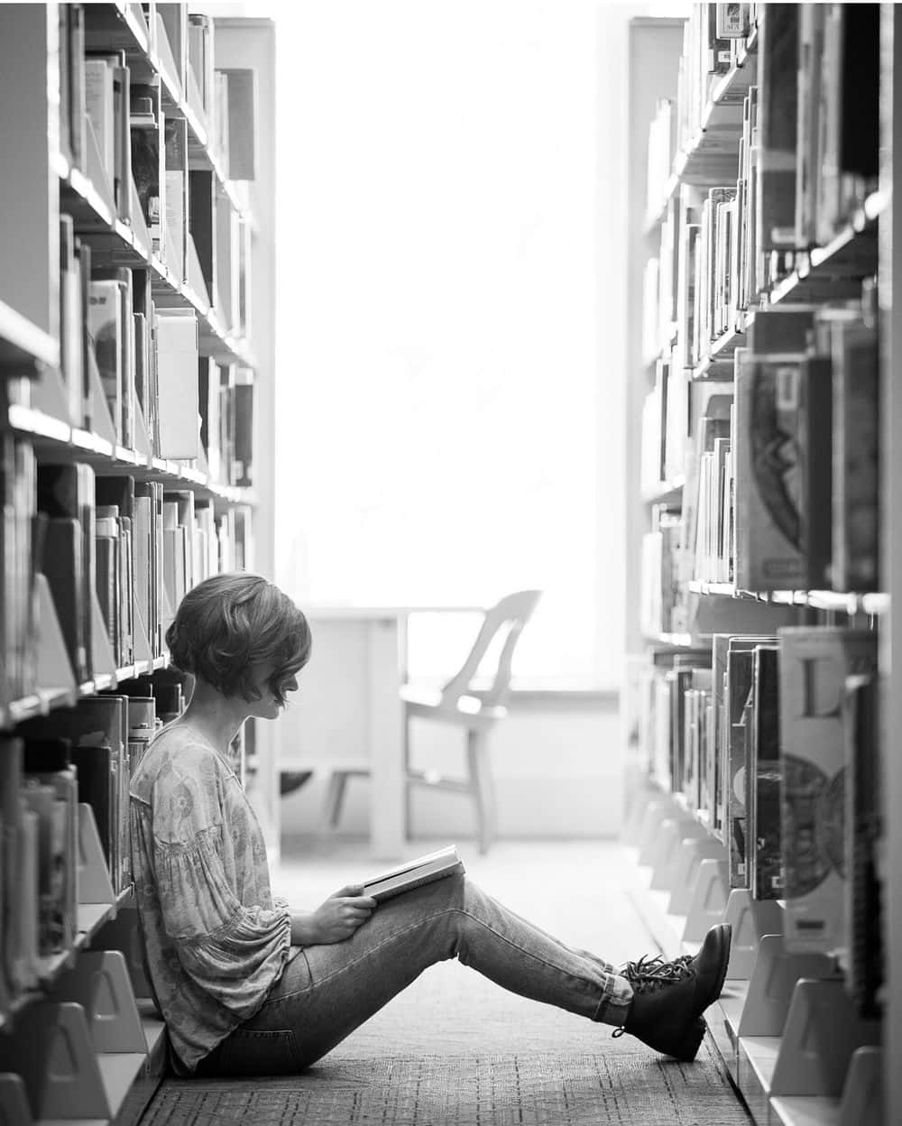 Library 桌布 壁紙 — 一名女性在陽光普照的圖書館走廊中，盤腿坐在高聳的書架之間閱讀一本書。