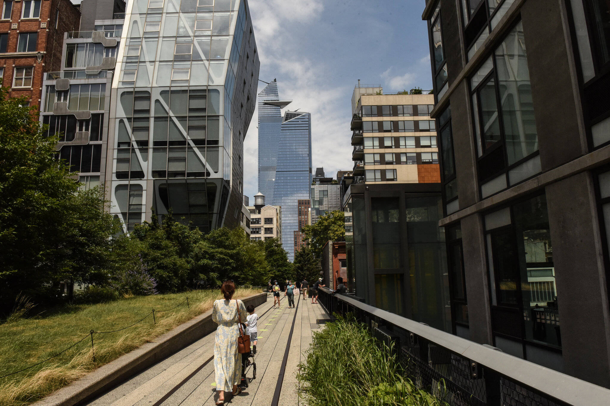 New York 桌布 壁紙 — 一名女性在陽光普照的高架人行道上推著嬰兒車，兩旁是現代玻璃建築和綠化，頭頂是藍天。