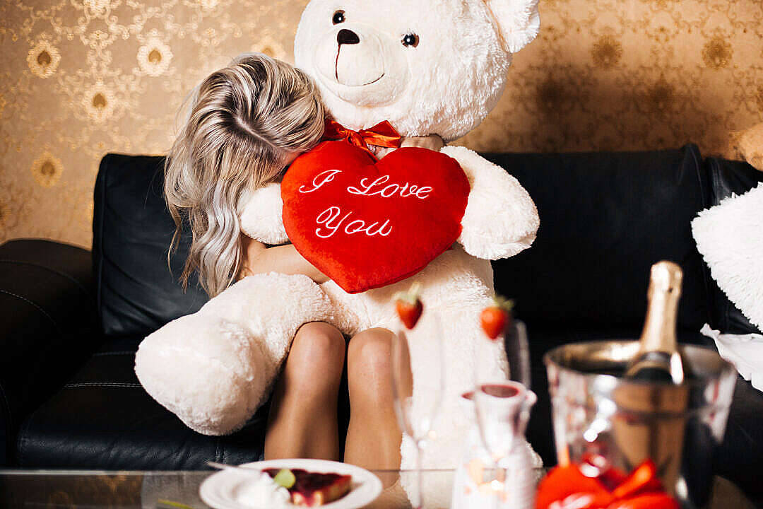 Cute Teddy Bear 桌布 壁紙 — 一名女性坐在黑色皮革沙發上，擁抱一隻拿著寫有「I Love You」的紅色心形枕頭的巨型白色泰迪熊。