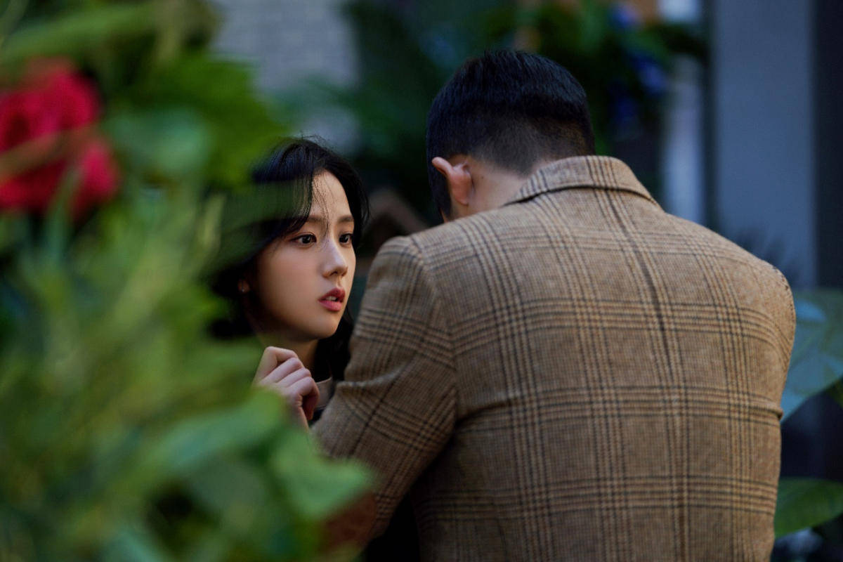 drama 桌布 壁紙 — 一名女性從茂密綠植後方觀看一名穿著格紋外套的男性，焦點在紅色花朵上。