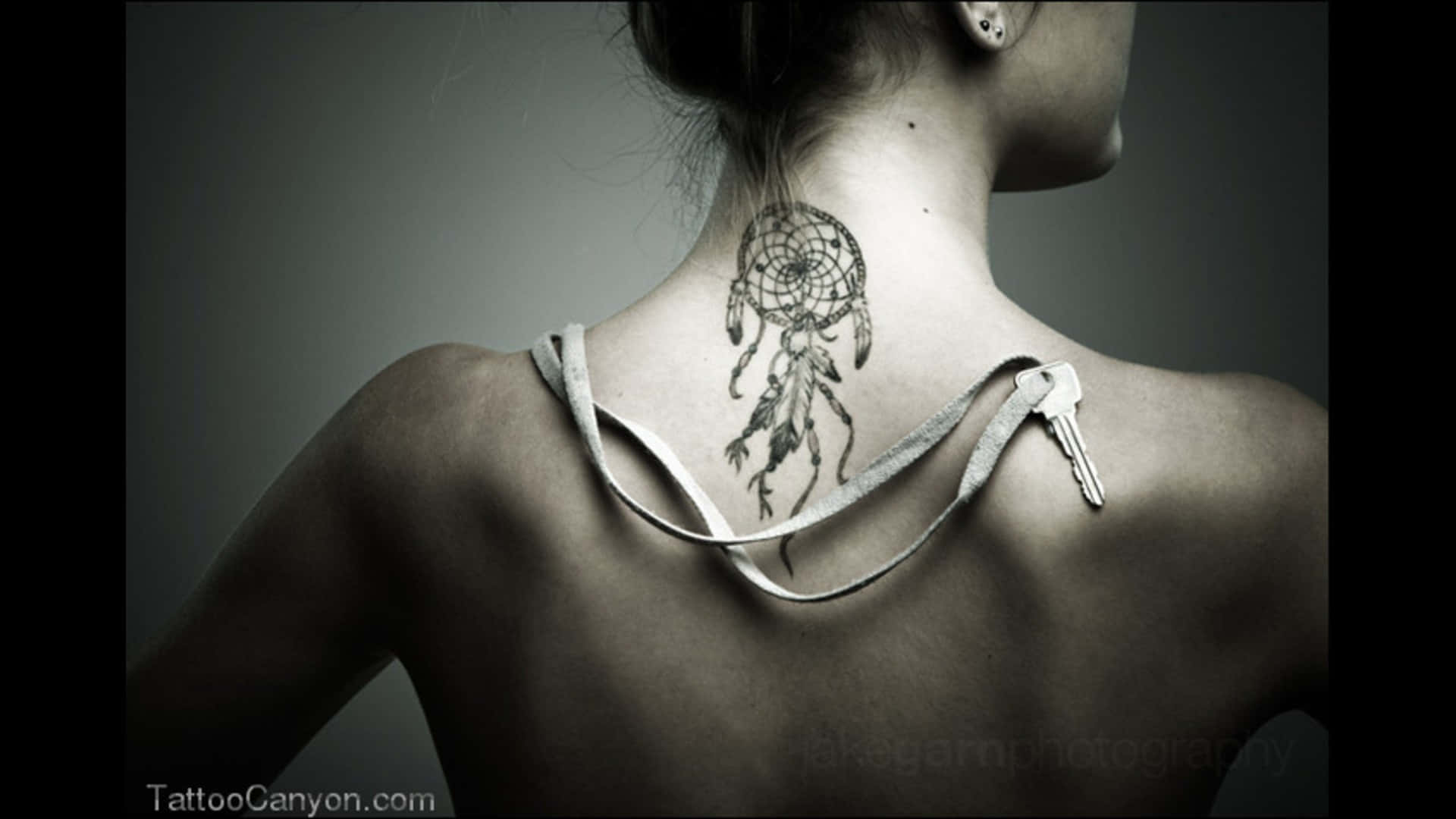 Tattoo 桌布 壁紙 — 一名女性的背部展示了一個捕夢網刺青，一條鑰匙從她的肩帶上垂下，背景為柔和的灰色。