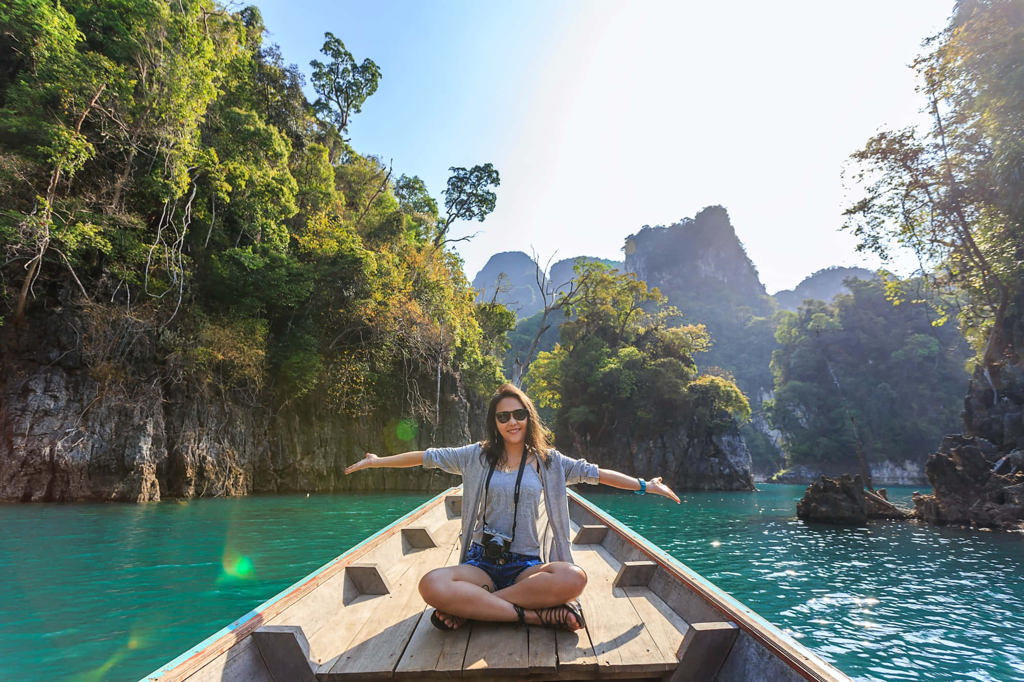 Traveling 桌布 壁紙 — 一名女性盤腿坐在木船上，船漂浮在碧綠色的水面上，周圍是鬱鬱蔥蔥的綠色懸崖，陽光普照。