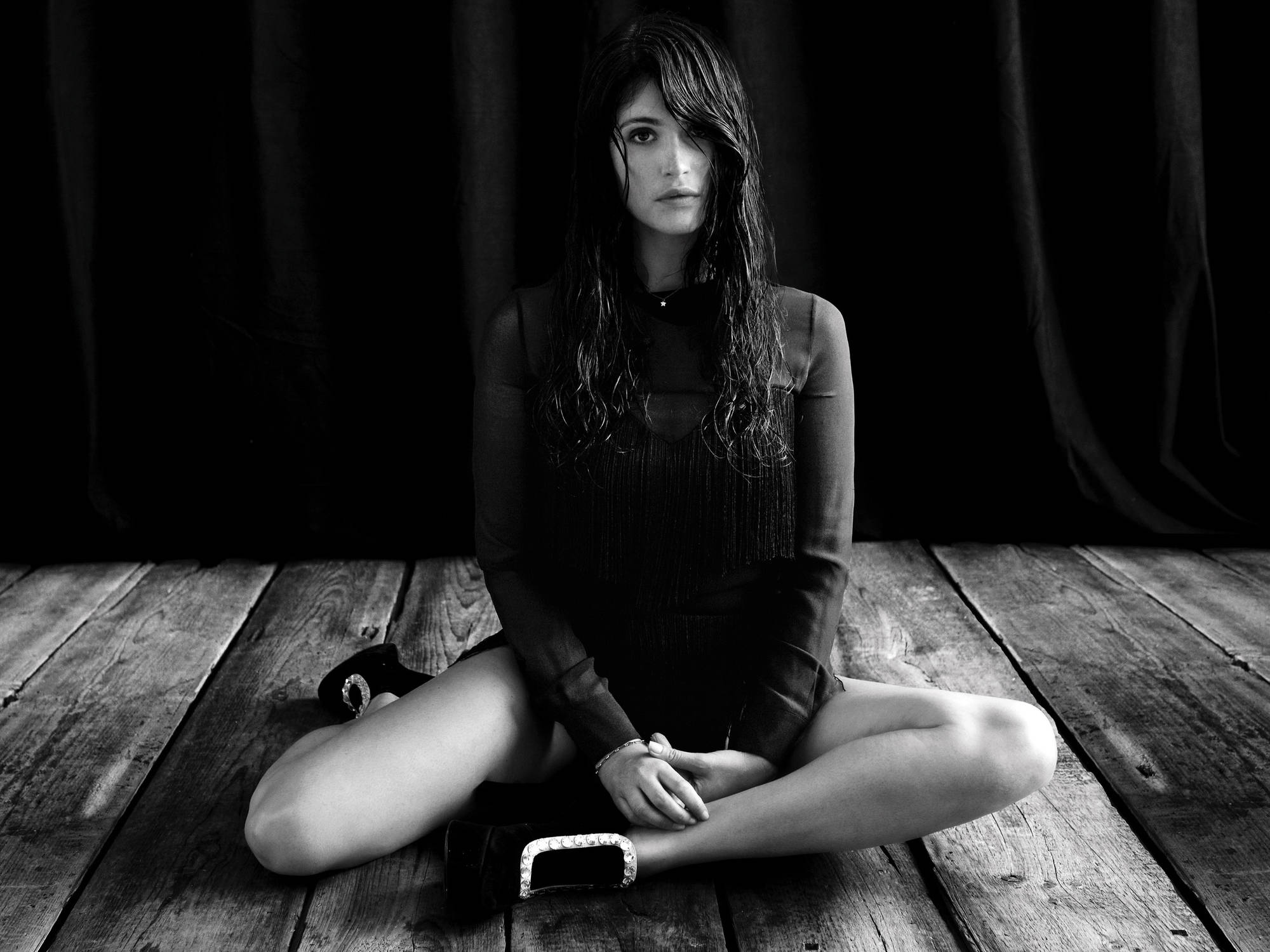 Gemma Arterton 桌布 壁紙 — 一名女性盤腿坐在破舊的木地板上，背景是深色窗簾，形成鮮明的黑白肖像。