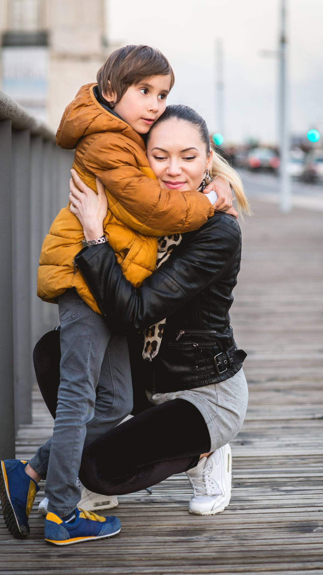 Hug 桌布 壁紙 — 一名女性跪在木製步道上，擁抱著一個穿著蓬鬆橙色外套的孩子，兩人凝視著鏡頭。