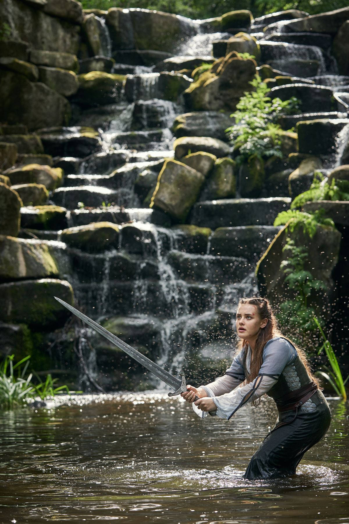Katherine Langford 桌布 壁紙 — 一名女戰士涉水而行，手持劍，身後瀑布在長滿苔蘚的岩石上傾瀉而下。