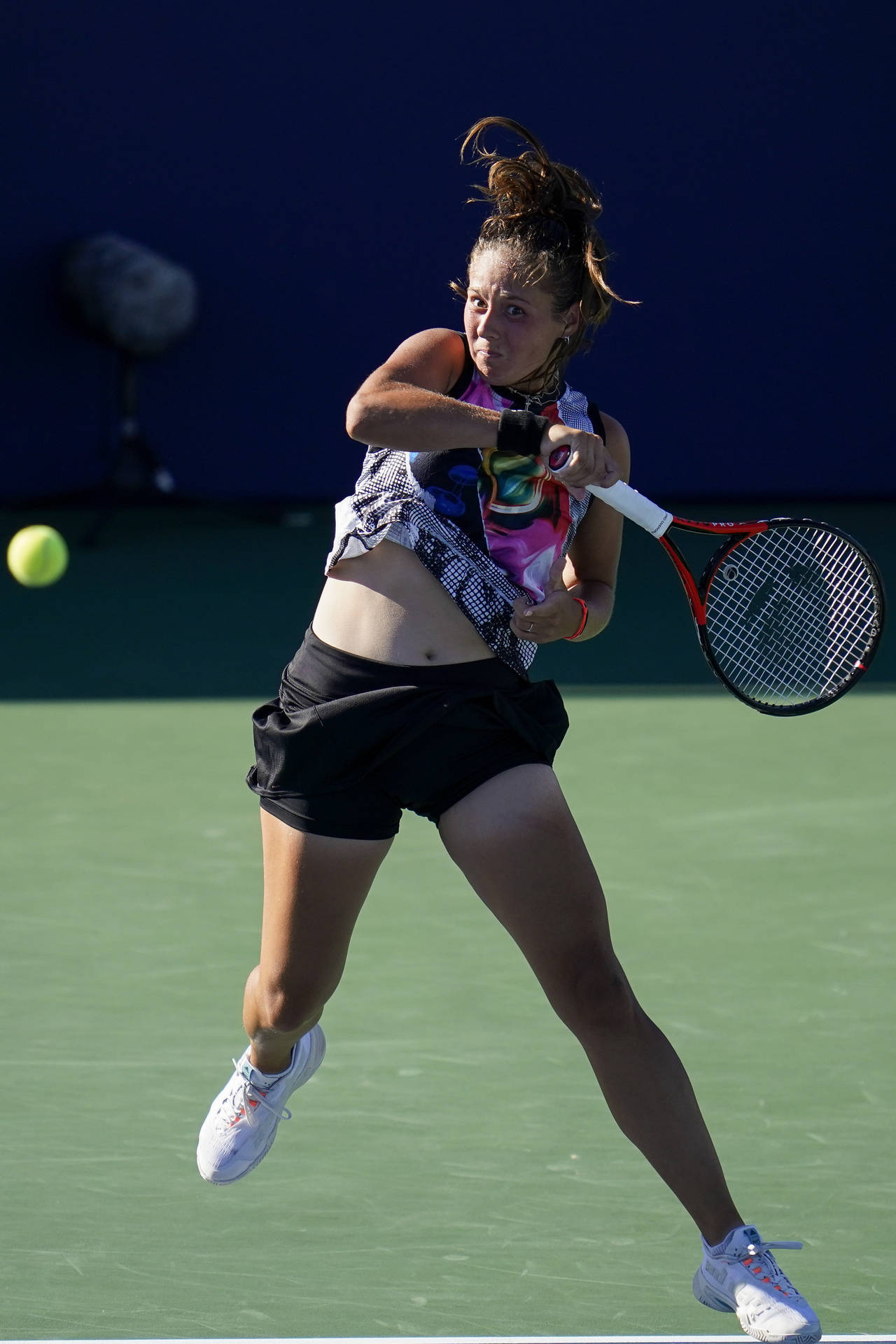 Athletic 桌布 壁紙 — 一名女網球運動員在半空中跳躍，揮動球拍擊向綠色球場上的一個黃色球，在明亮的日光下。