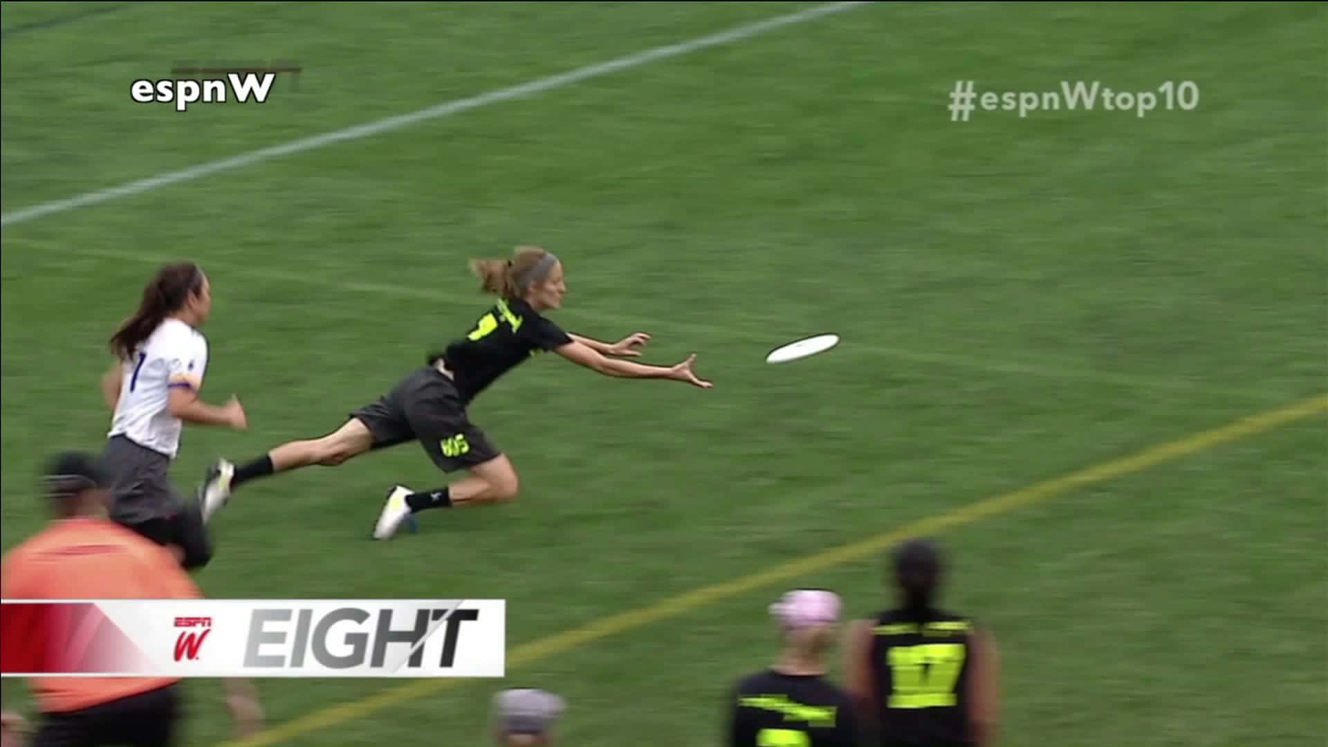 Frisbee 桌布 壁紙 — 一名女運動員在草地上躍起，伸手接住飛行的飛盤，隊友和對手在旁觀看。