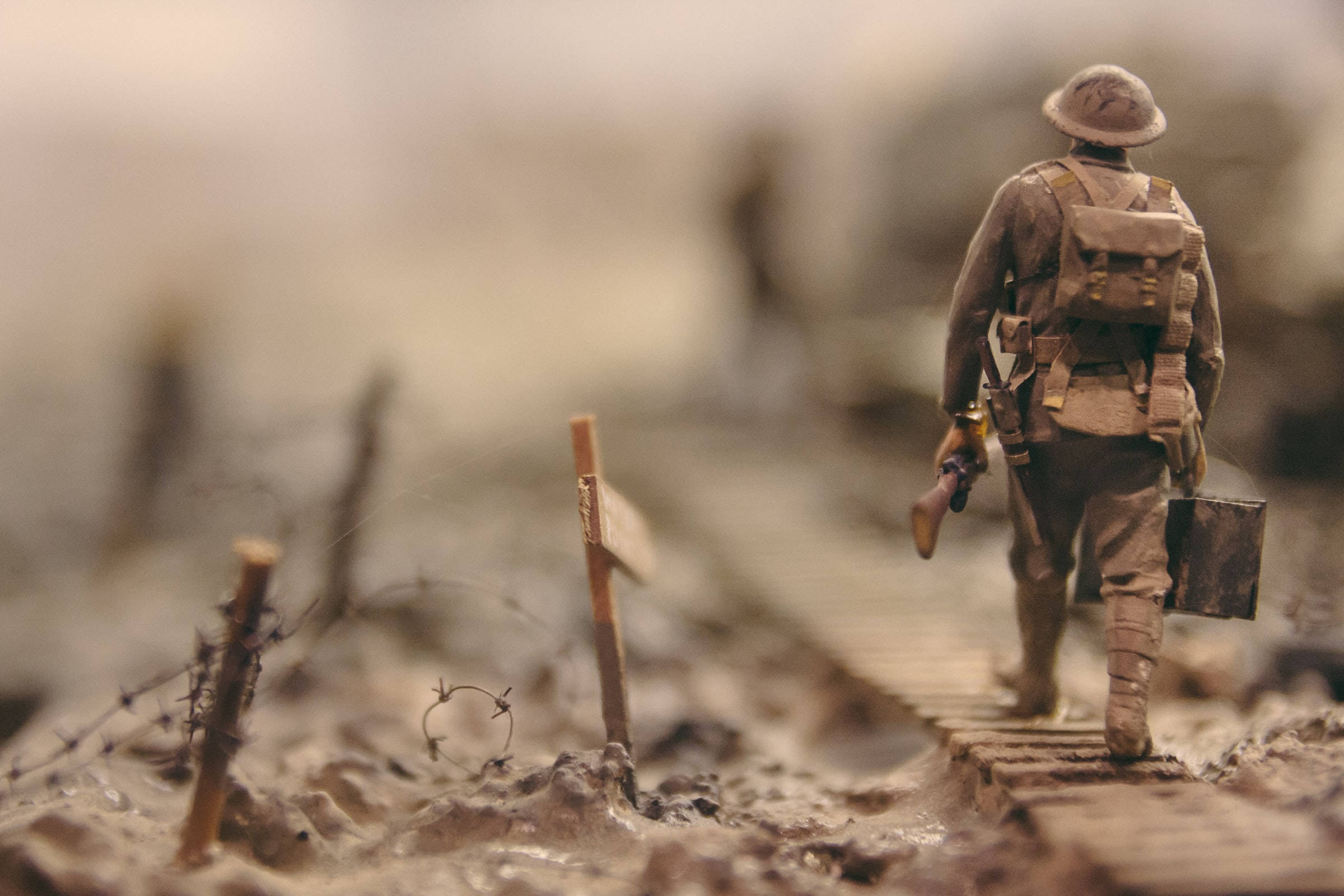 Remembrance 桌布 壁紙 — 一名孤獨的士兵身穿泥濘的戰壕裝備，在木板上走過一個被戰爭摧毀的戰場，遠處有鐵絲網和模糊的人影。