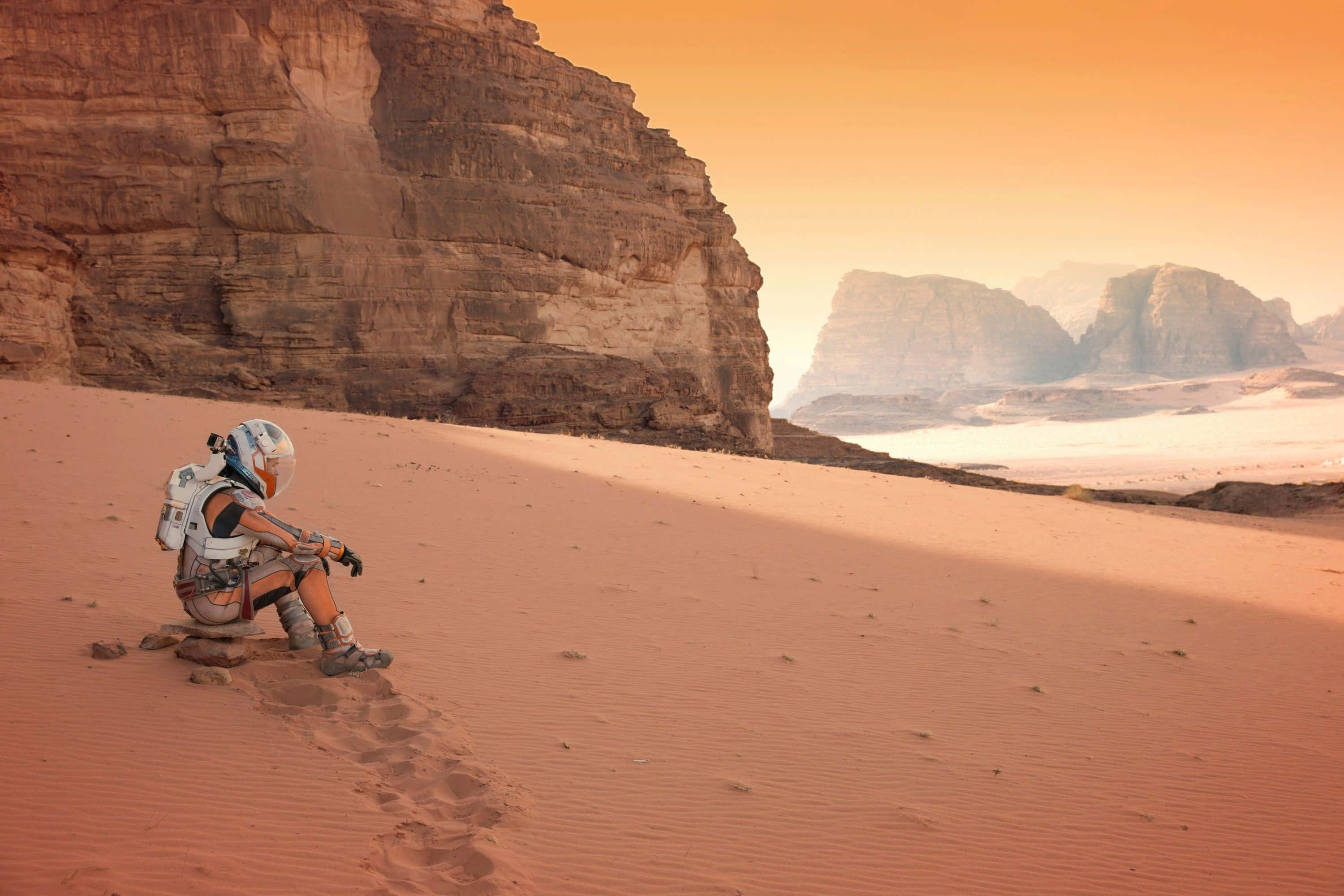The Martian 桌布 壁紙 — 一名孤獨的太空人戴著白色頭盔，坐在紅色沙丘上，溫暖的橙色天空下，遠處有高聳的岩石 formations。
