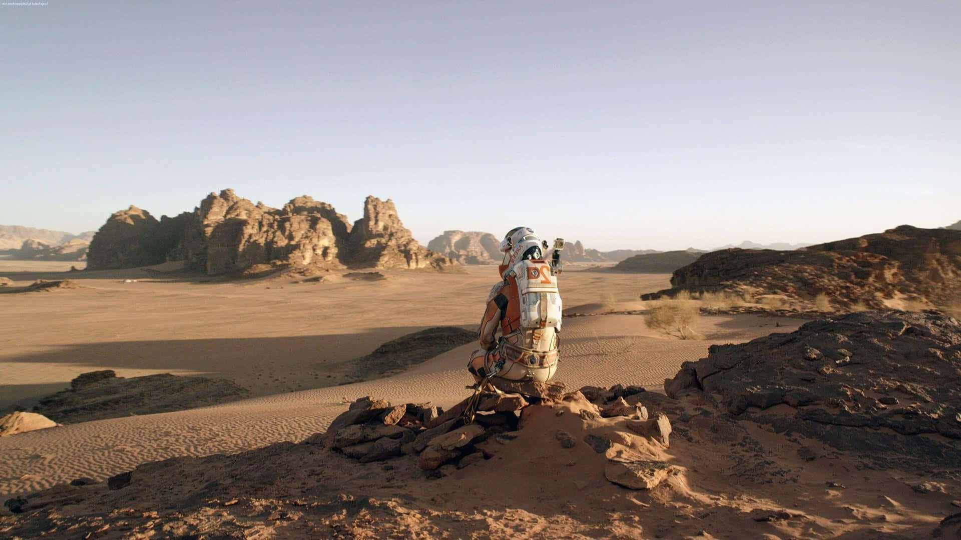 The Martian 桌布 壁紙 — 一名孤獨的太空人身穿白色太空衣，跪在岩石沙漠山脊上，俯瞰廣闊的沙丘和遠處的岩石地貌，天空晴朗。