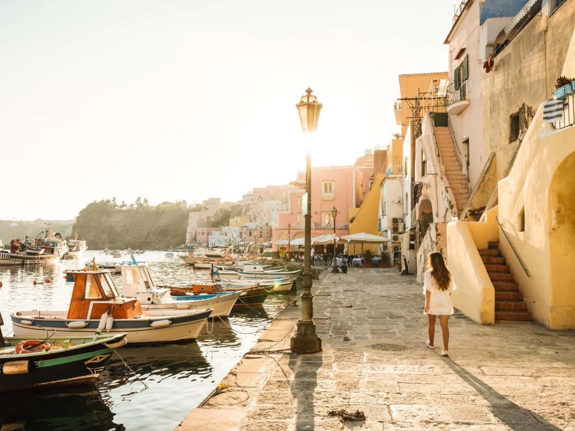 Italian Island 桌布 壁紙 — 一名孤獨的女子在陽光照耀的鵝卵石碼頭上行走，經過色彩繽紛的建築物和停泊的漁船，正值黃金時刻。