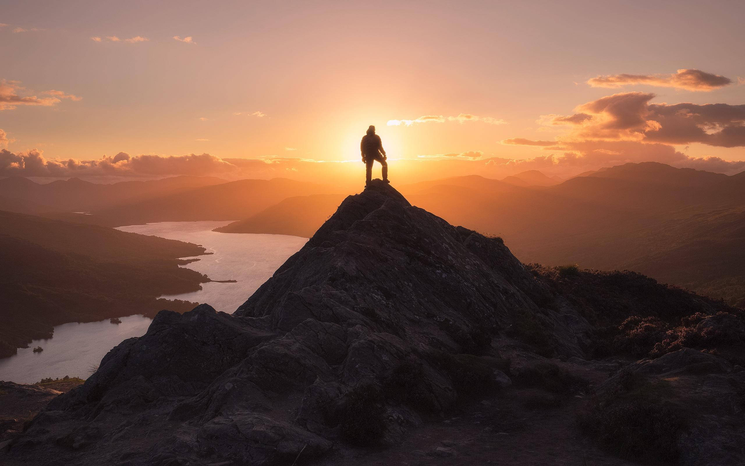 Victory 桌布 壁紙 — 一名孤獨的徒步旅行者在崎嶇的山頂上剪影，日落時分俯瞰蜿蜒的湖泊和遠山。