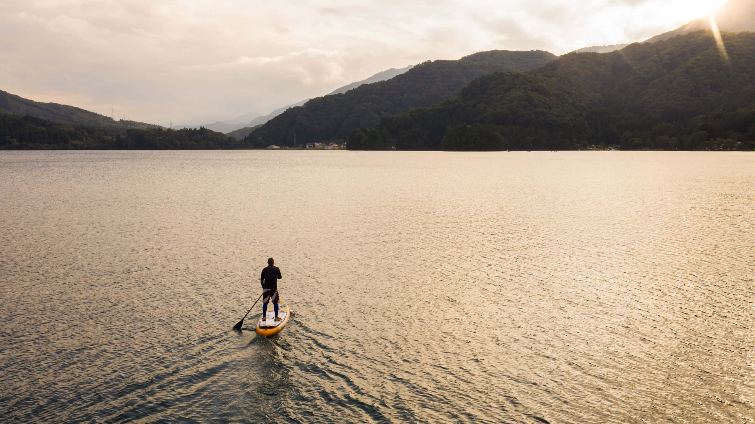 Paddleboarding 桌布 壁紙 — 一名孤獨的槳板運動者在金色日落時分滑過寧靜的山間湖泊，遠處是森林覆蓋的山丘。