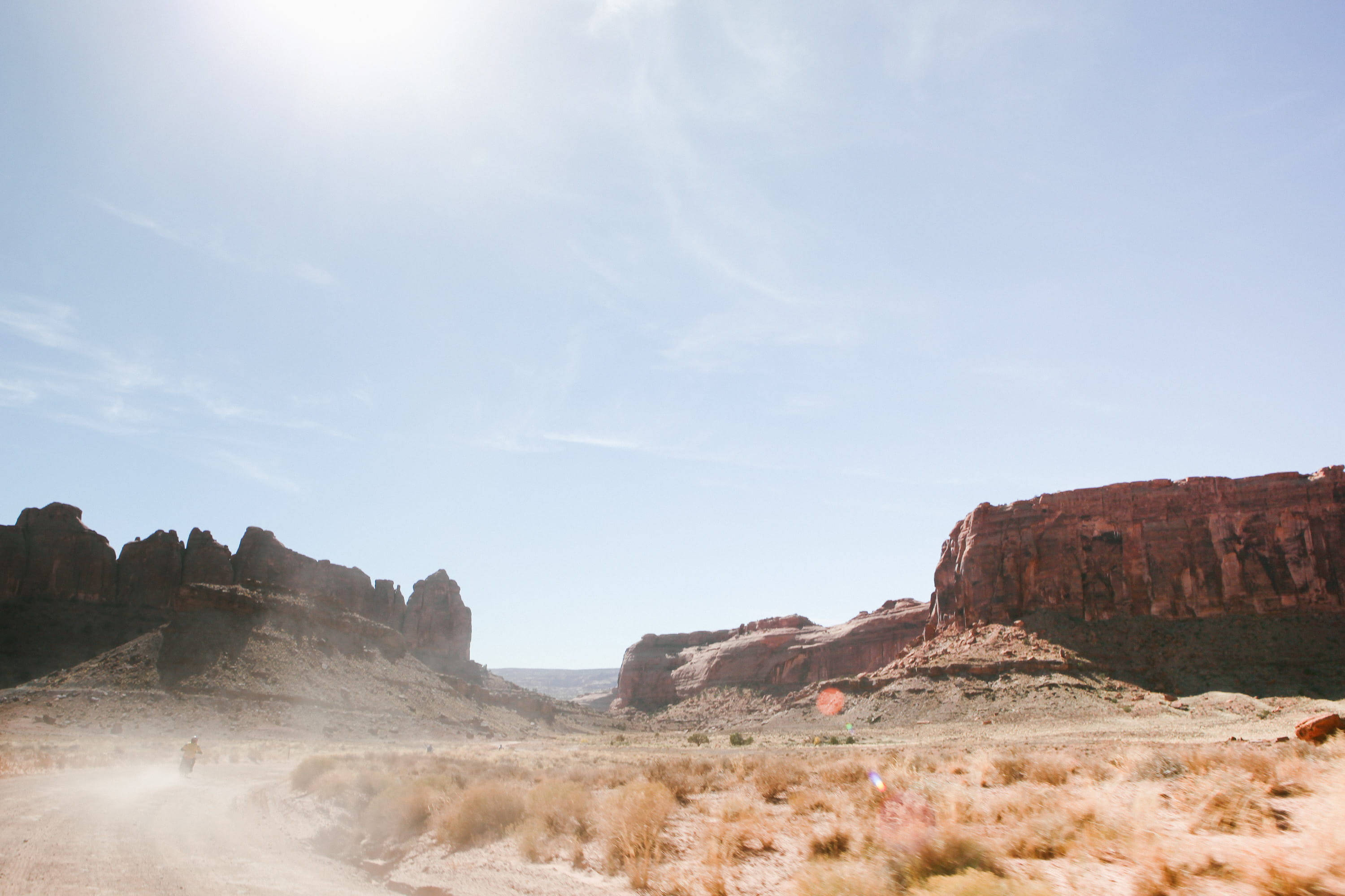 Canyonlands National Park 桌布 壁紙 — 一名孤獨的機車騎士在晴朗藍天下，於兩旁高聳紅色岩層的炎熱沙漠小徑上揚起塵土。