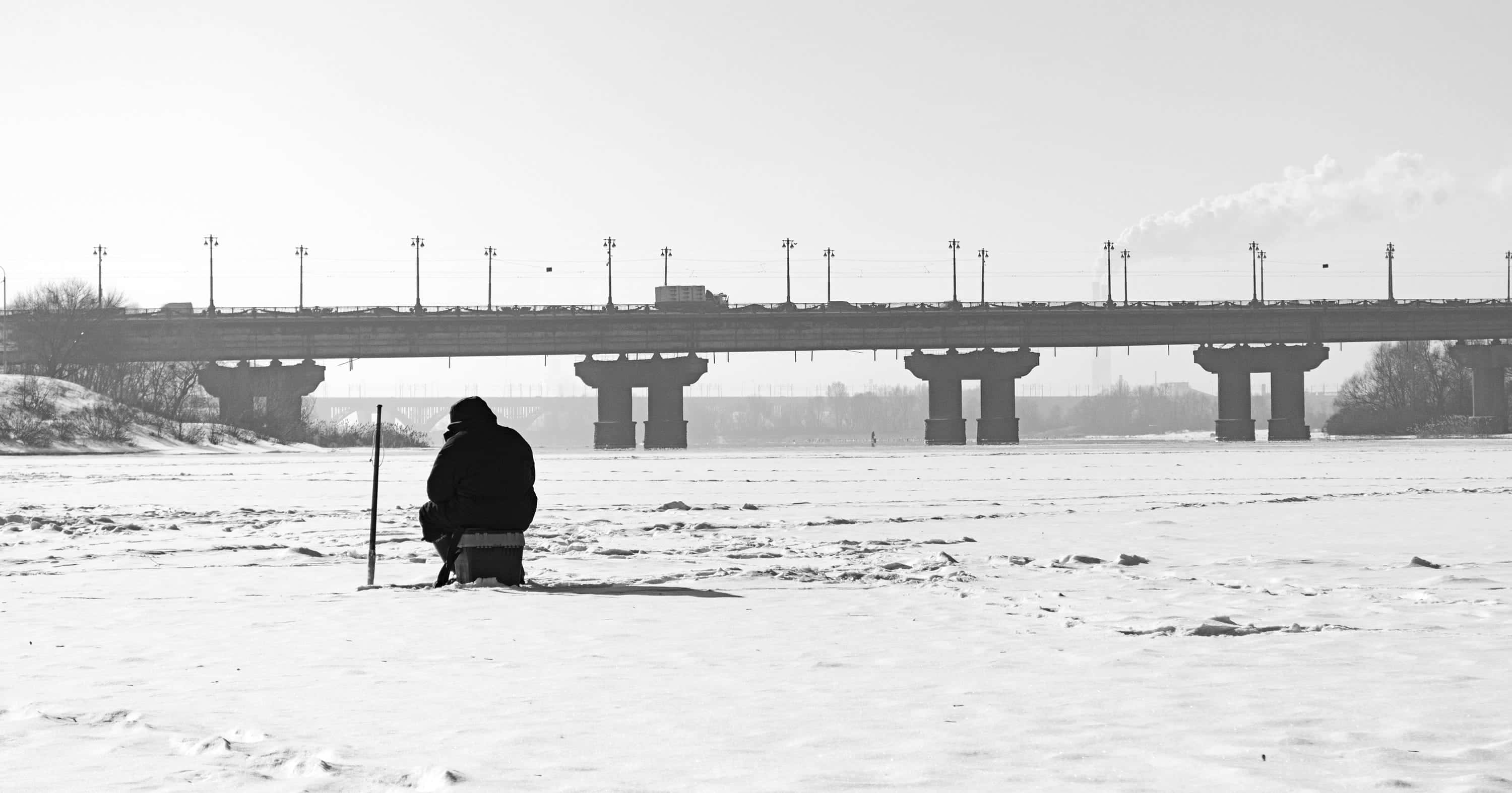Ice Fishing 桌布 壁紙 — 一名孤獨的漁夫坐在結冰的河上，廣闊的冬日天空下，遠處有一座橋橫跨冰封的河面。