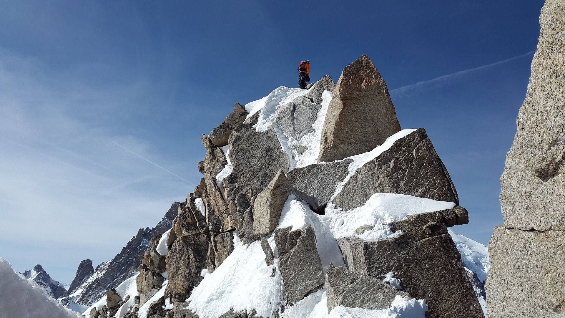 Climbing 桌布 壁紙 — 一名孤獨的登山者身穿紅色裝備，站在白雪覆蓋的花崗岩山峰上，天空湛藍，遠處山脊連綿。