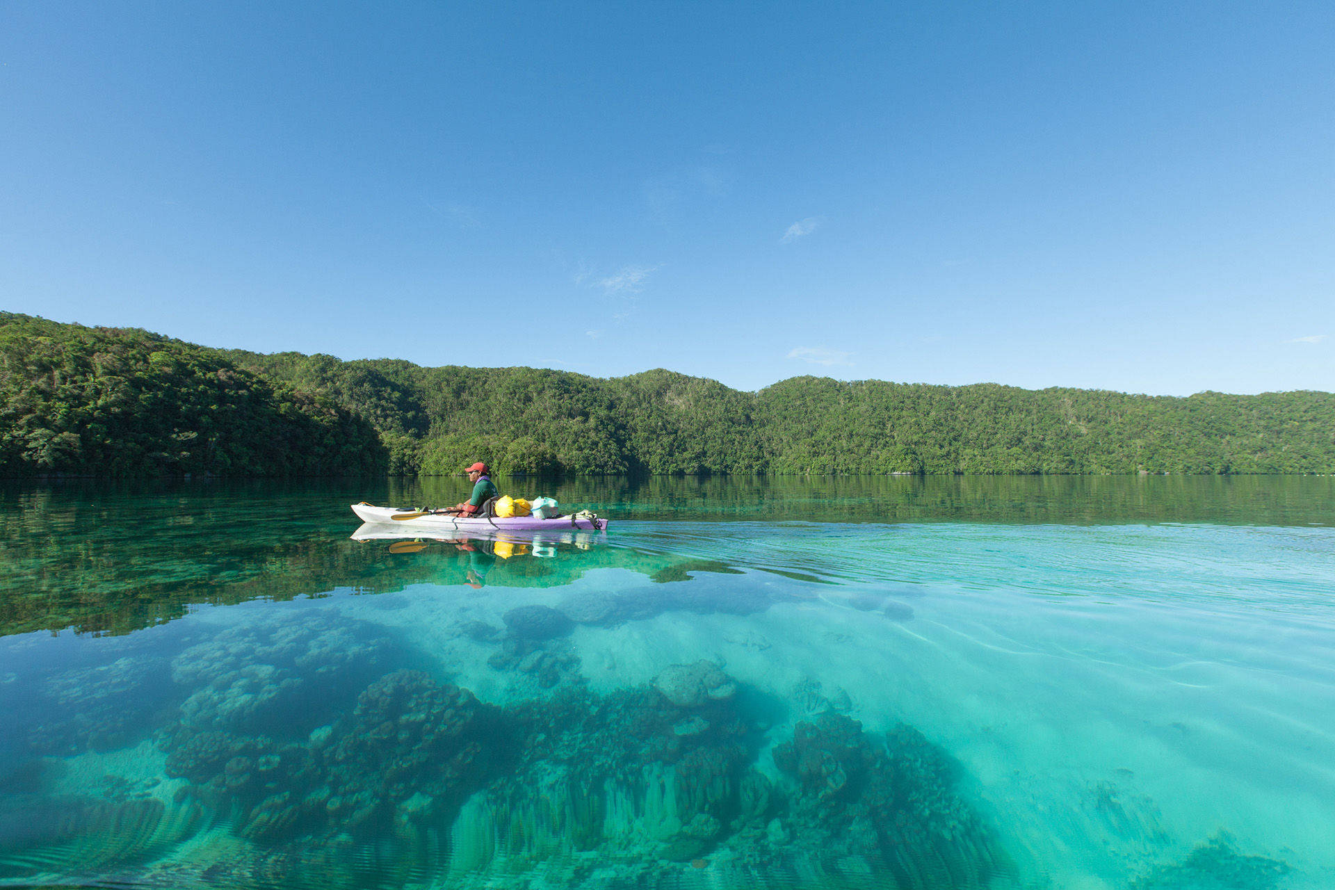 Micronesia 桌布 壁紙 — 一名孤獨的皮划艇運動員在清澈碧綠的水面上滑行，露出下方充滿活力的珊瑚礁，而鬱鬱蔥蔥的綠色山丘在廣闊的藍天下聳立。