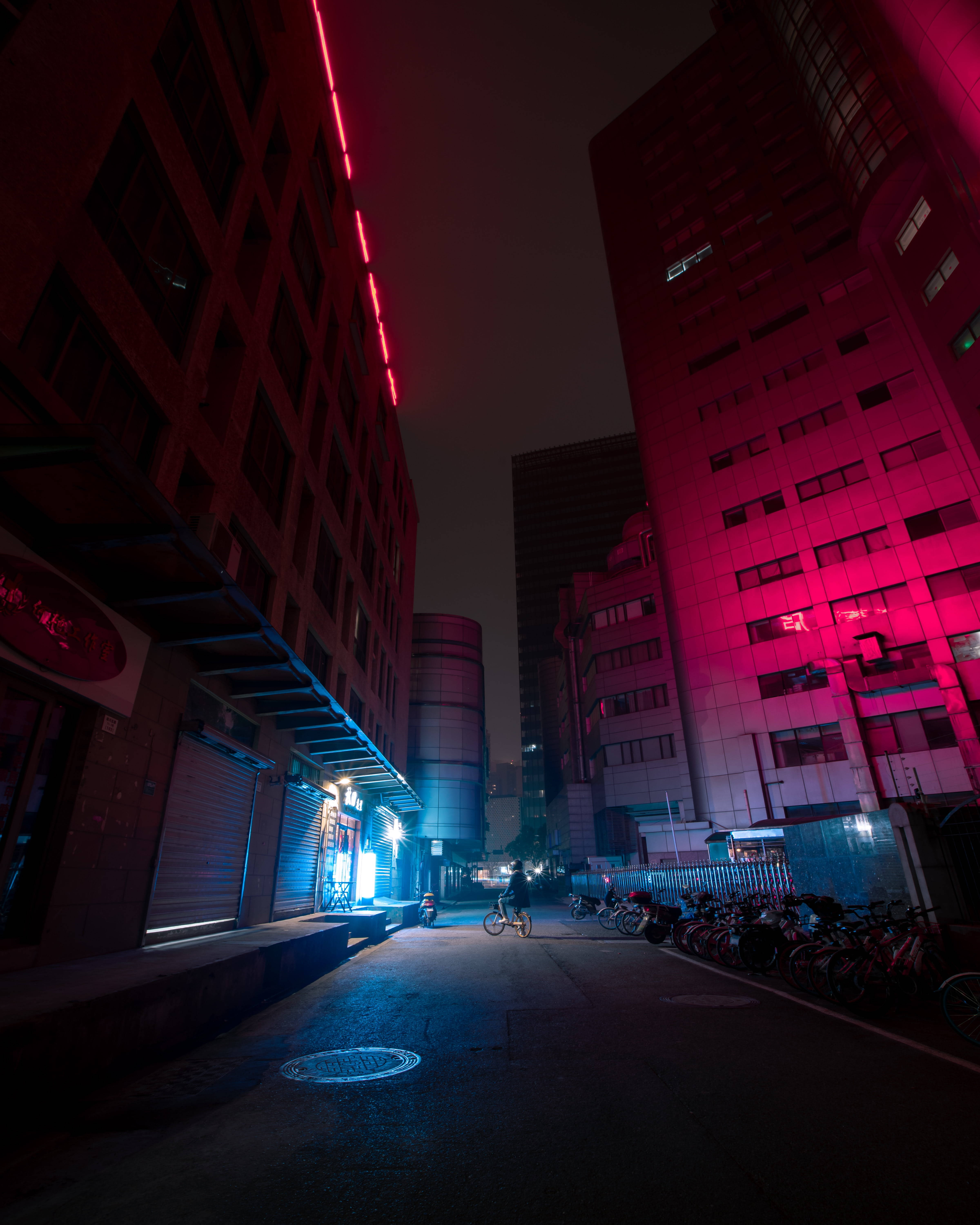 Dark Neon 桌布 壁紙 — 一名孤獨的自行車騎士在夜間沿著狹窄的城市小巷騎行，被高樓大廈鮮豔的紅色和藍色霓虹燈照亮。