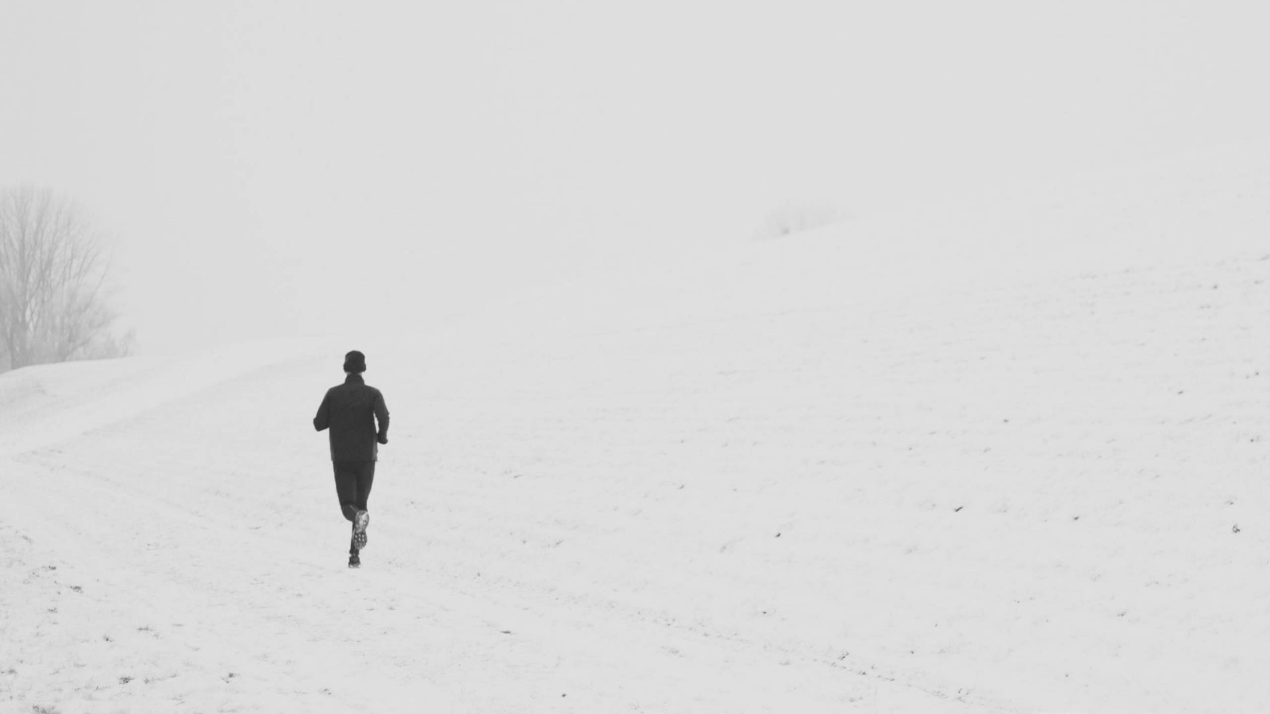 Running 桌布 壁紙 — 一名孤獨的跑者身穿深色裝備，在灰濛濛的冬日天空下，穿過廣闊的雪地向上衝刺。