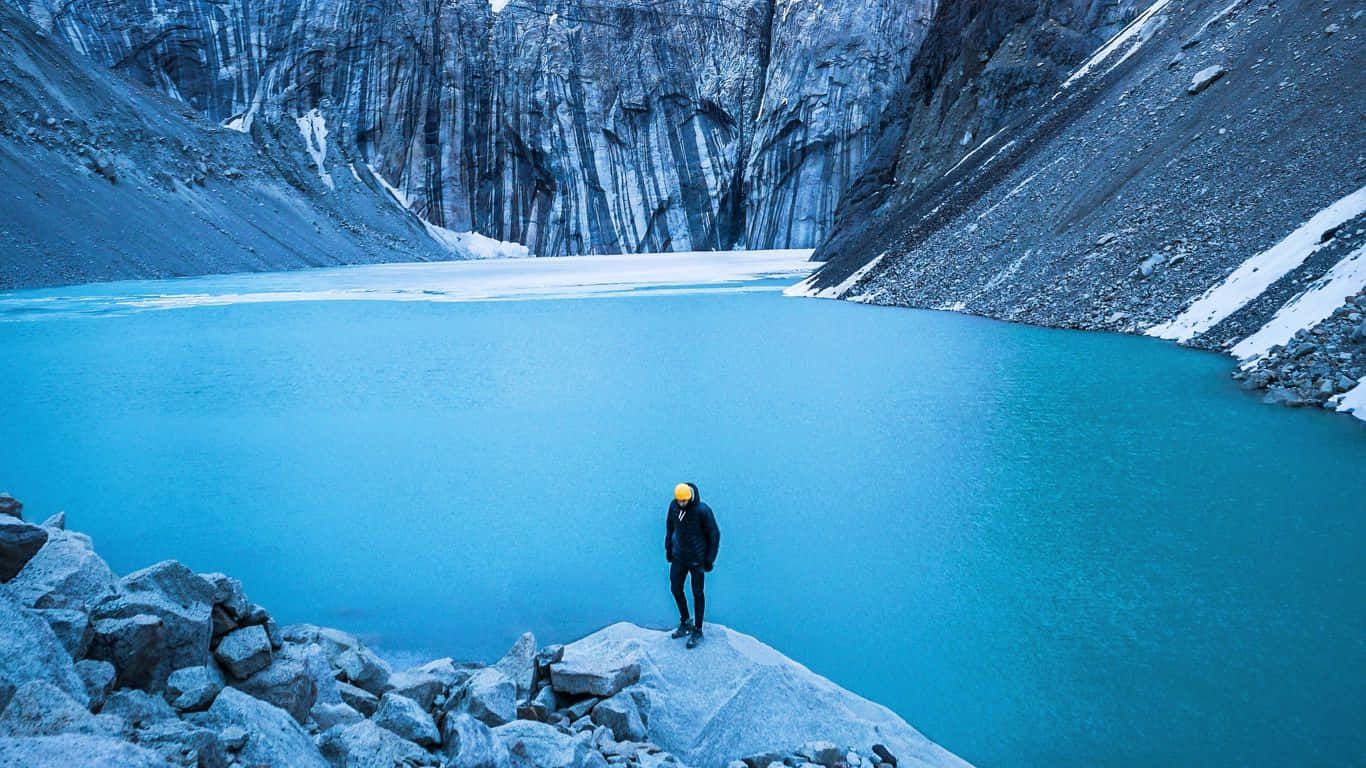 Traveler 桌布 壁紙 — 一名孤獨的遠足者戴著黃色頭盔，站在岩石露頭上，俯瞰著一個被雪塵覆蓋的懸崖環繞的令人驚嘆的綠松石冰川湖。