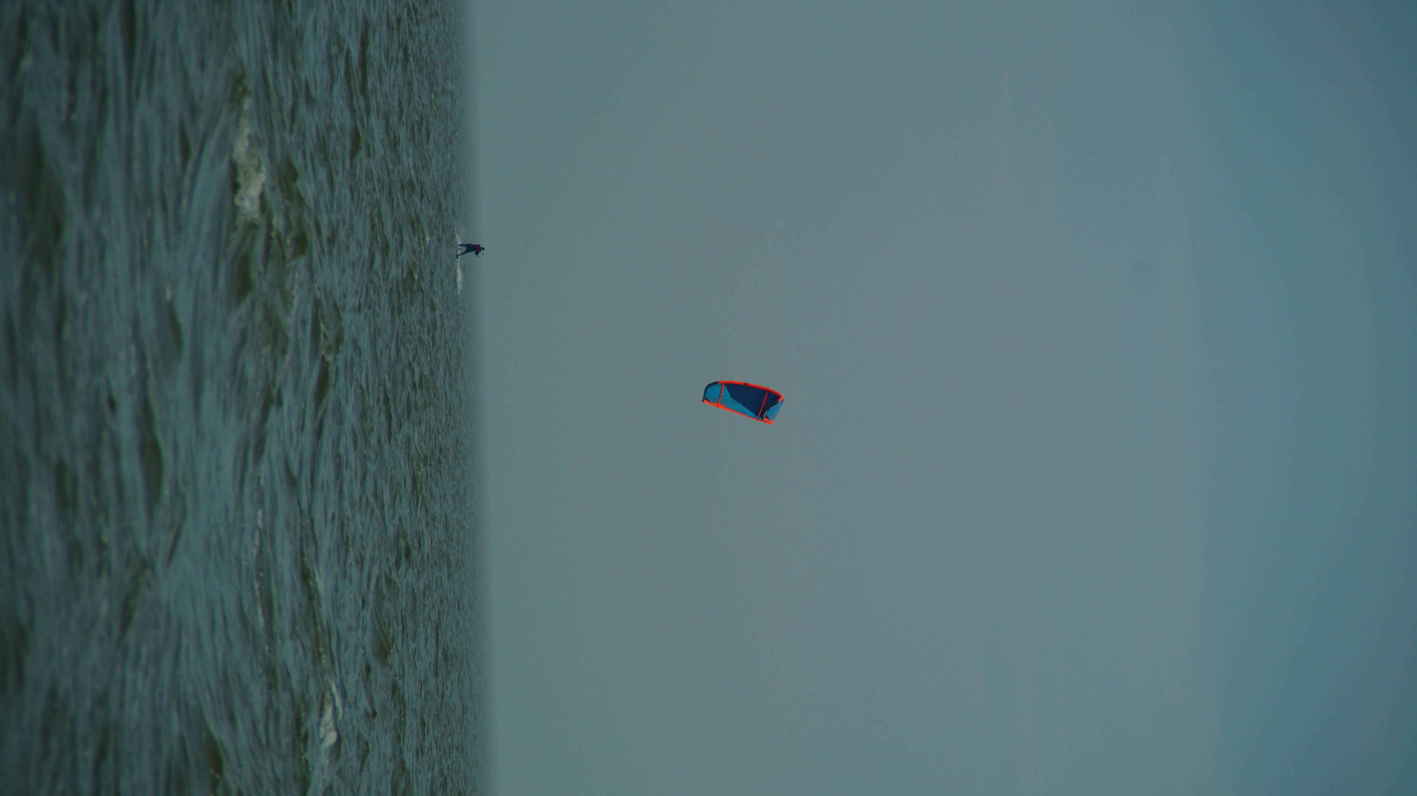 Windsurfing 桌布 壁紙 — 一名孤獨的風箏衝浪者在廣闊陰天之下，滑過波濤洶湧的藍綠色水面，鮮紅與藍色的風箏高高飛揚。