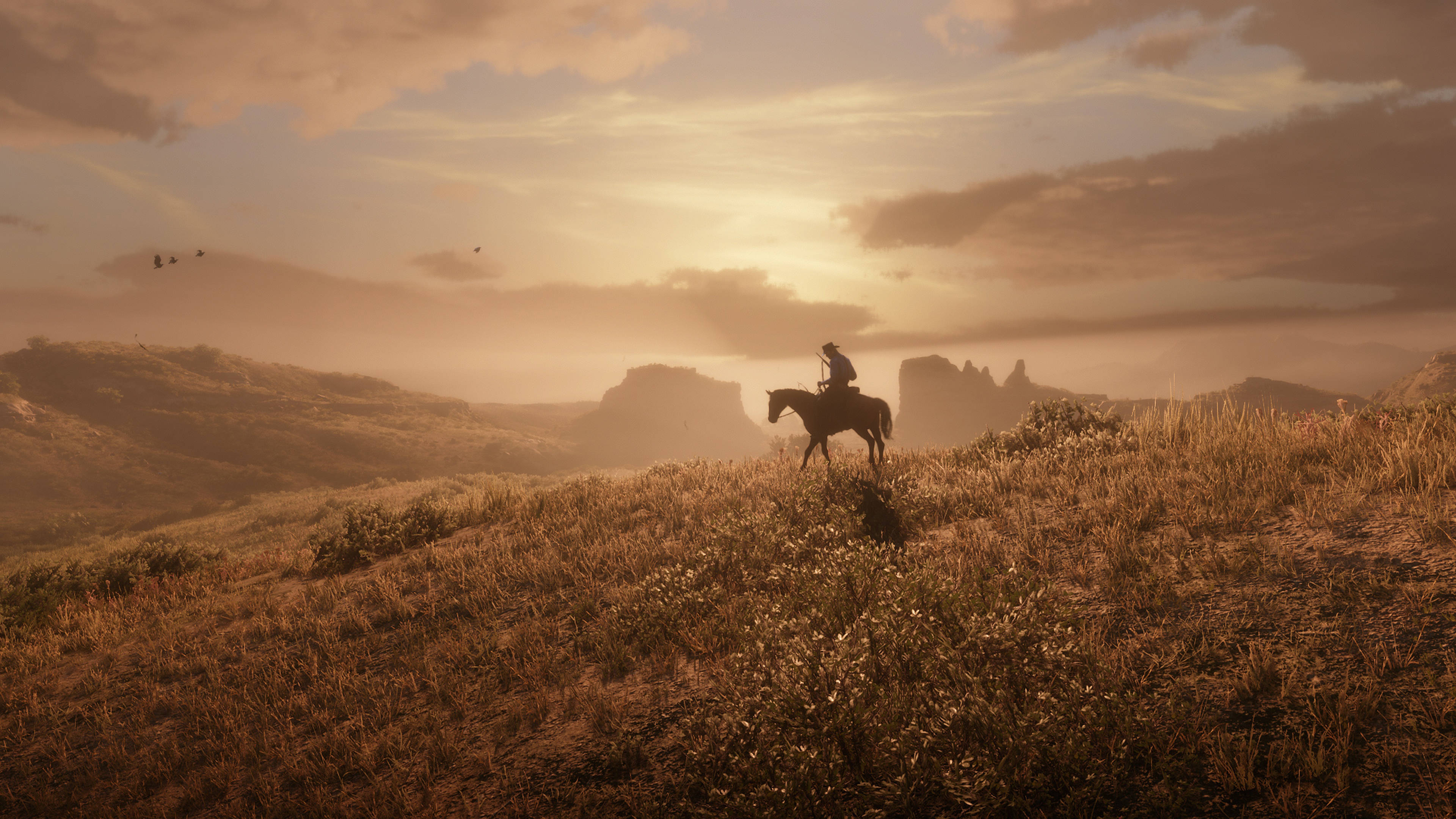Red Dead Redemtion 2 桌布 壁紙 — 一名孤獨的騎手騎在馬上，在崎嶇的沙漠山丘和遠處岩石地貌上，以金色日落為背景形成剪影。