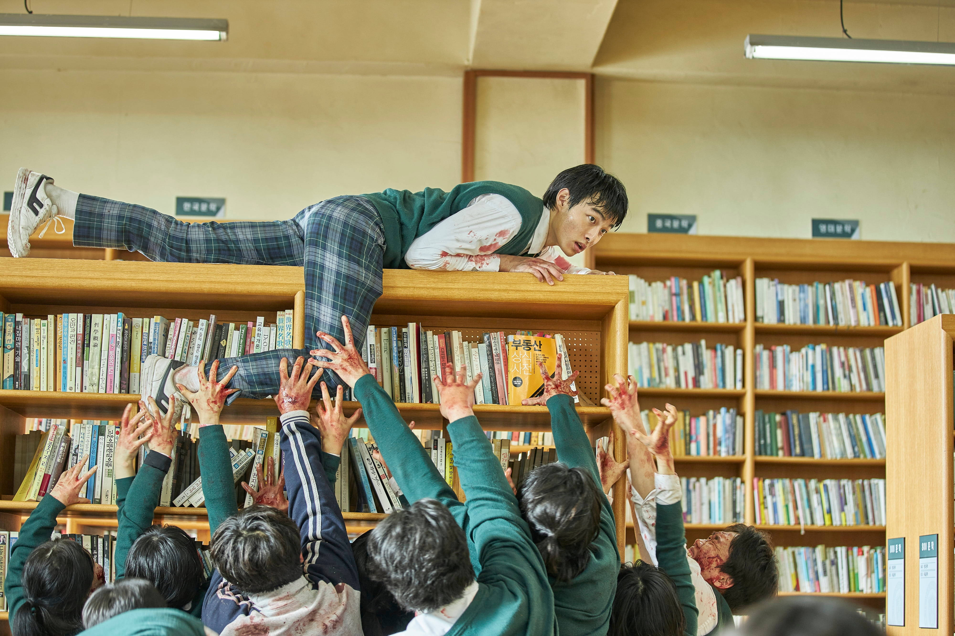 殭屍動圖 桌布 壁紙 — 一名學生滿身鮮血躺在圖書館書架上，而下面的同學則伸出沾滿鮮血的手，營造出緊張的恐怖場景。