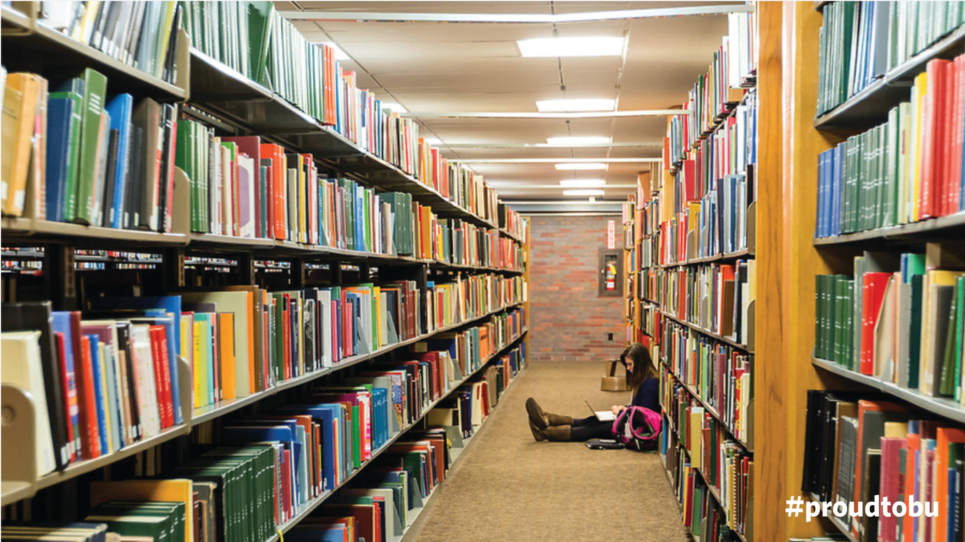 Library 桌布 壁紙 — 一名學生盤腿坐在圖書館地板上，周圍是螢光燈下充滿彩色書籍的高聳書架。