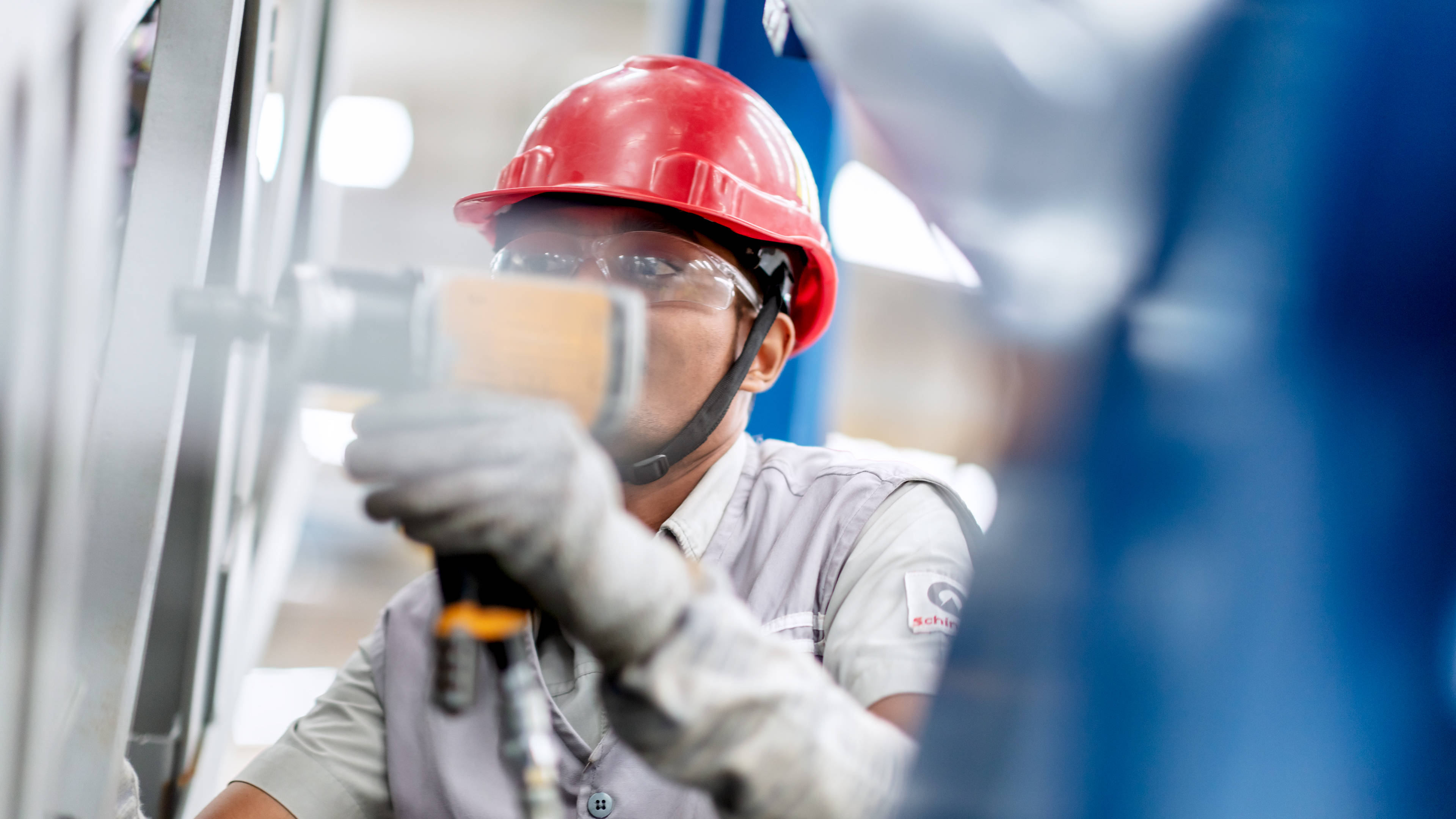 engineering 桌布 壁紙 — 一名專注的工業工人戴著紅色頭盔和護目鏡，在工廠環境中對金屬結構使用電動工具。
