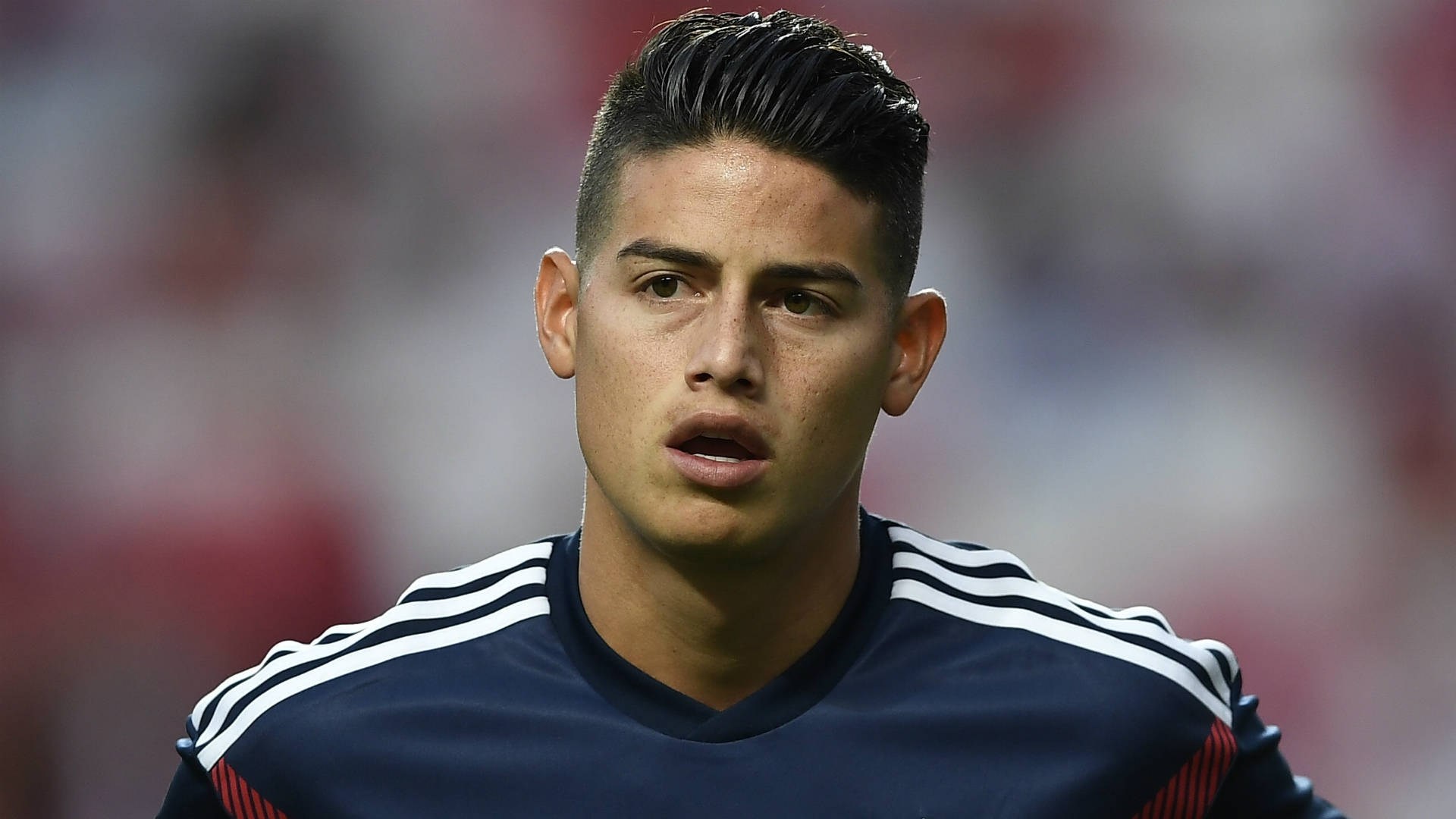 James Rodriguez 桌布 壁紙 — 一名專注的年輕足球員身穿帶有白色條紋的海軍藍球衣，在球場燈光下比賽時凝視著前方。