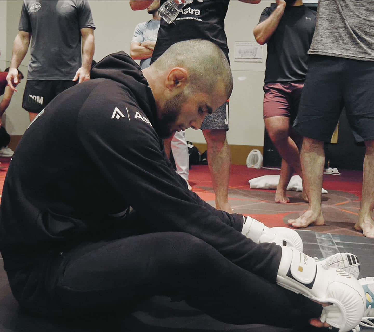 MMA 桌布 壁紙 — 一名專注的戰士穿著黑色裝備坐在墊子上繫白色手套，隊友們站在附近的訓練健身房中。