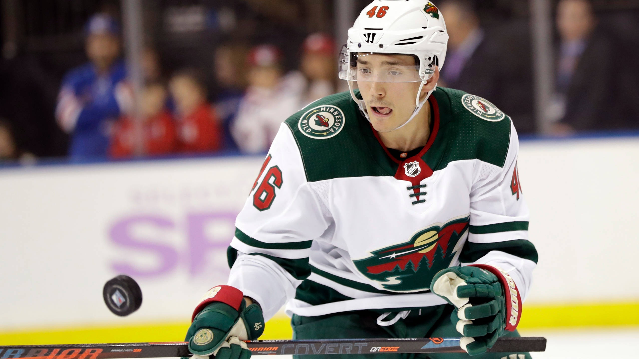 hockey 桌布 壁紙 — 一名專注的曲棍球運動員，身穿白色、綠色和紅色的制服，在比賽期間靠在冰上的球棍上。